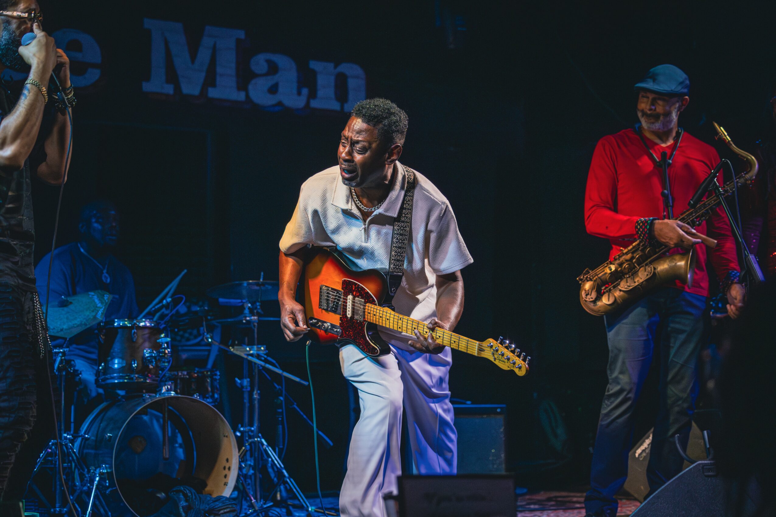 Musicians perform on stage with a drummer, guitarist, and saxophonist, under dark lighting and a blue backdrop.
