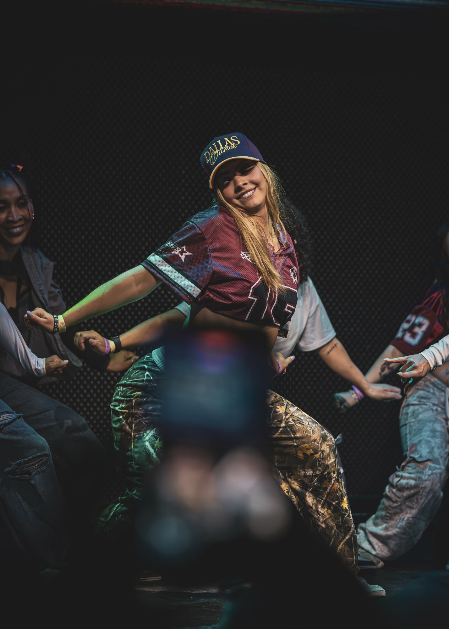 Group of people holding hands, smiling, with a woman in a baseball cap in the center, against a dark background.
