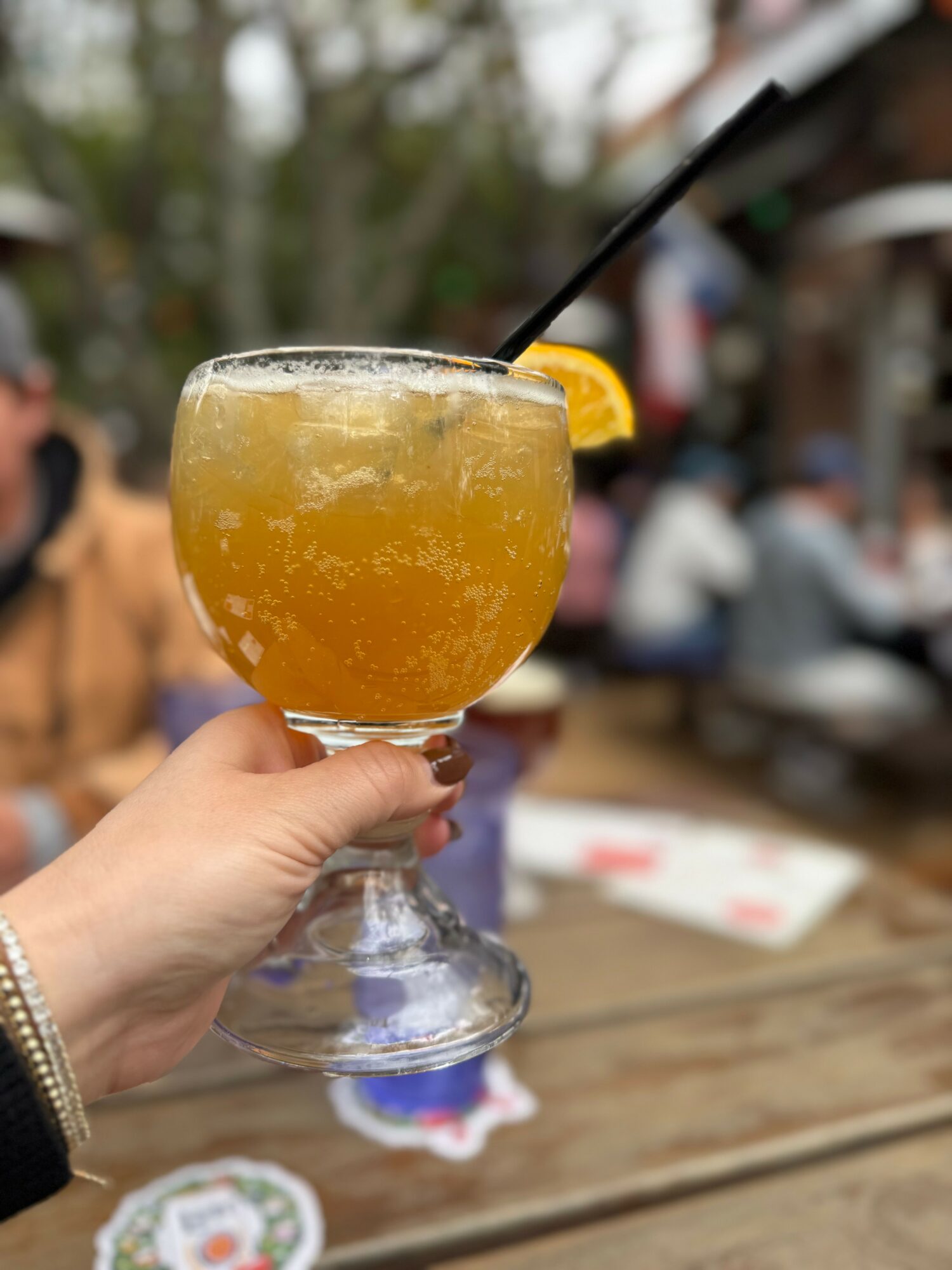 Hand holding a glass of yellow-orange beverage with a lemon slice, outdoor setting with blurred people and trees