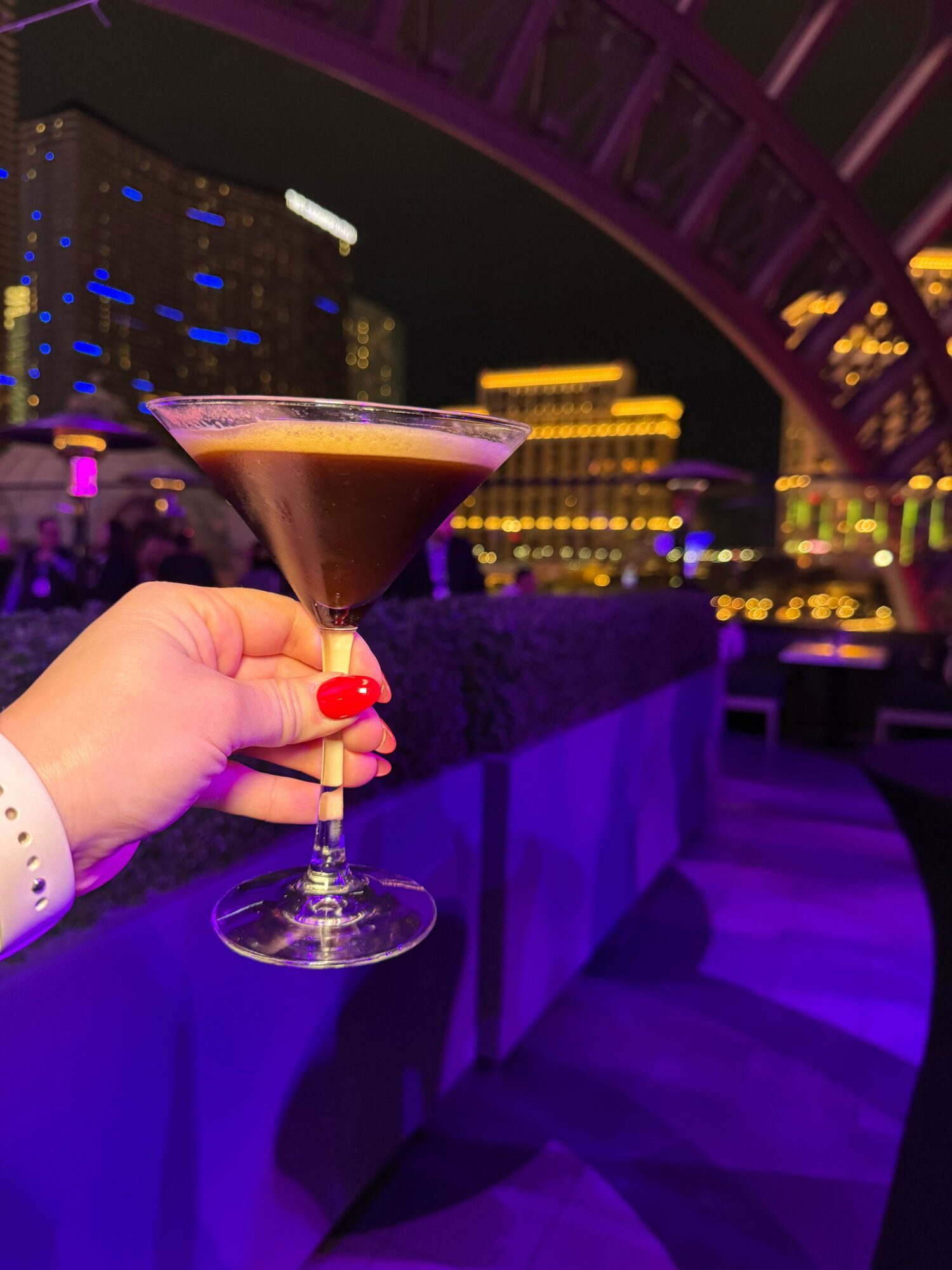 Hand holding a cocktail glass with a dark drink and a cherry garnish, against a city skyline at night, illuminated with purple lighting.