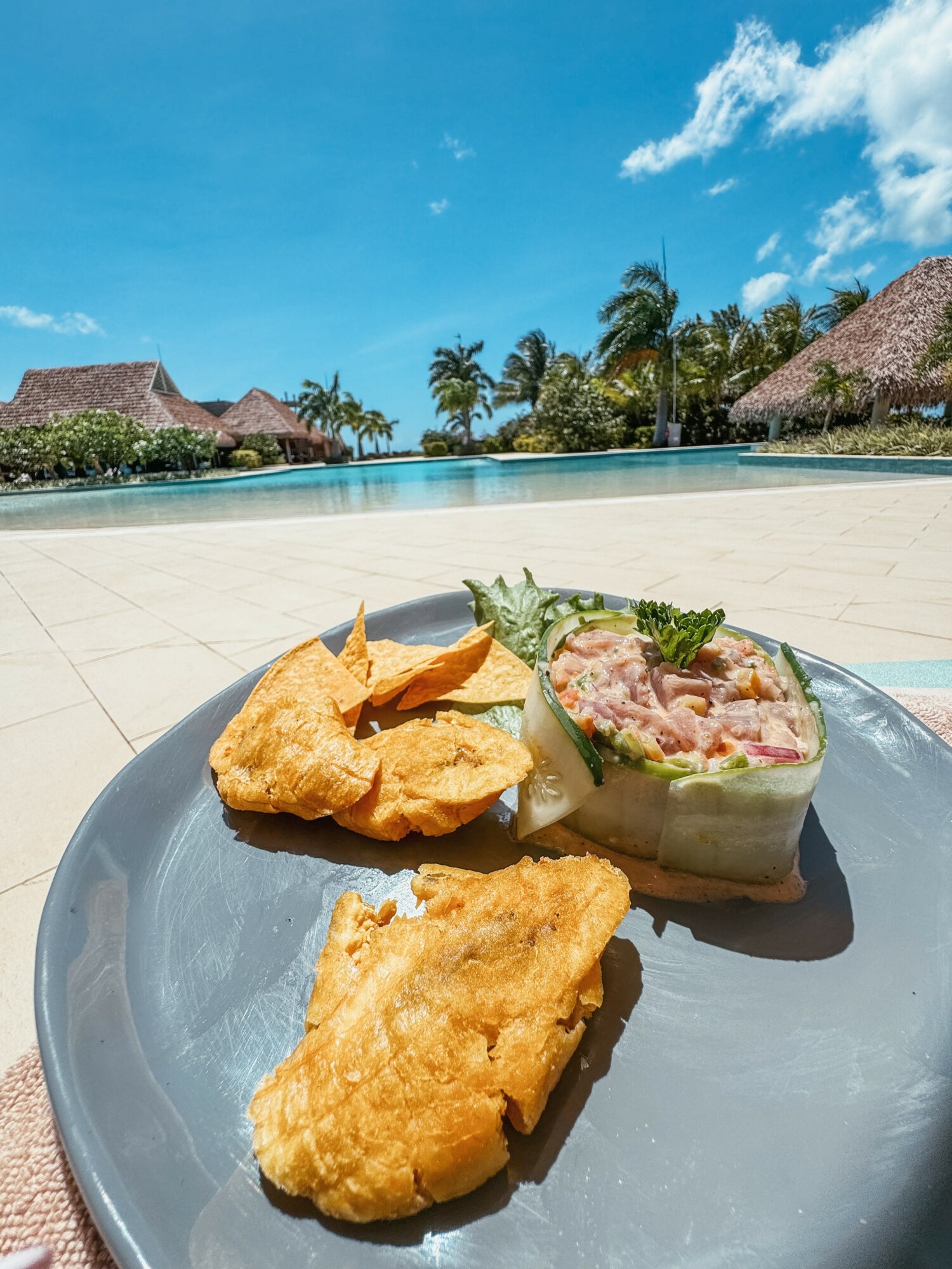 Plate with fried fish, chips, and a seafood salad on a poolside deck with huts and palm trees in the background.