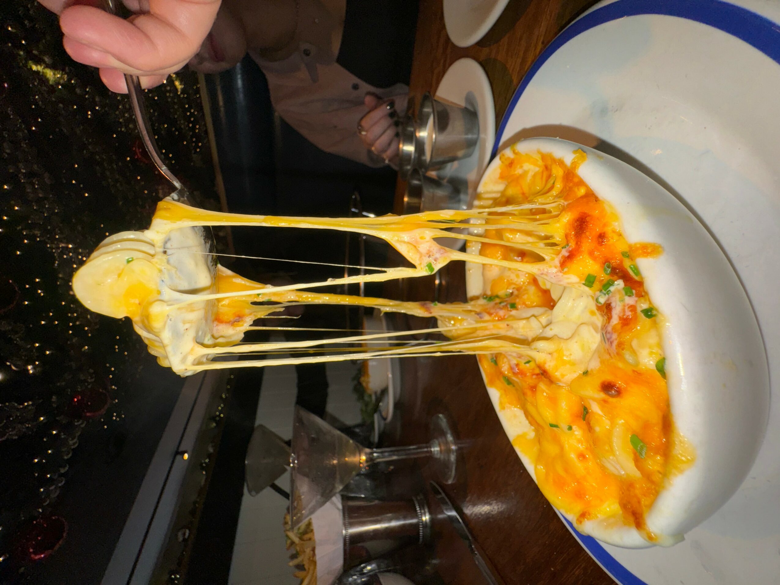 Person pouring melted cheese over a dish on a white plate with a blue rim, with a wooden table background.
