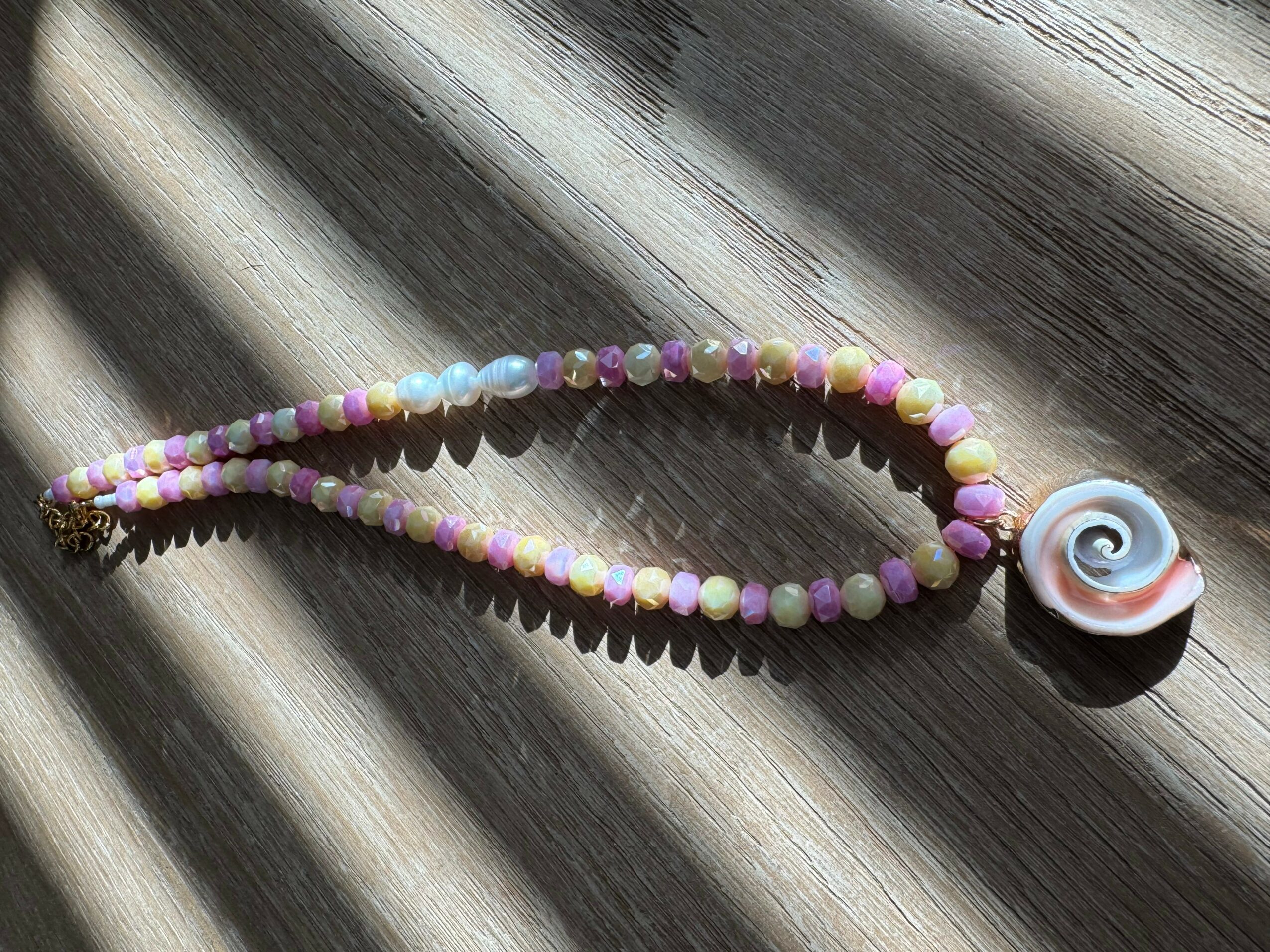 Beaded necklace with a spiral shell pendant on a wooden surface, sunlight casting shadows.