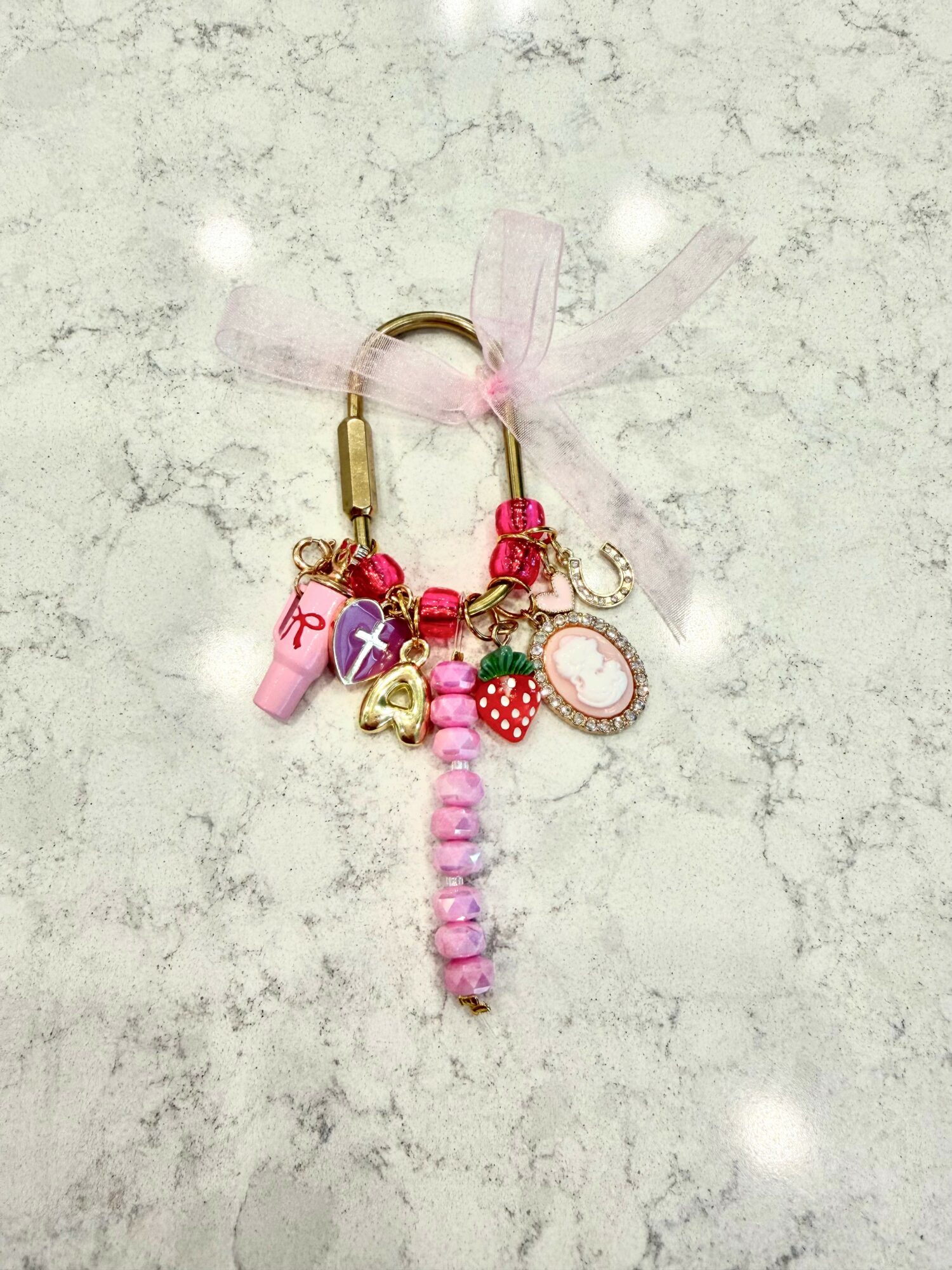 Charm keychain with pink beads, a pink bottle, a cupcake, and a heart-shaped mirror, on a light textured background.
