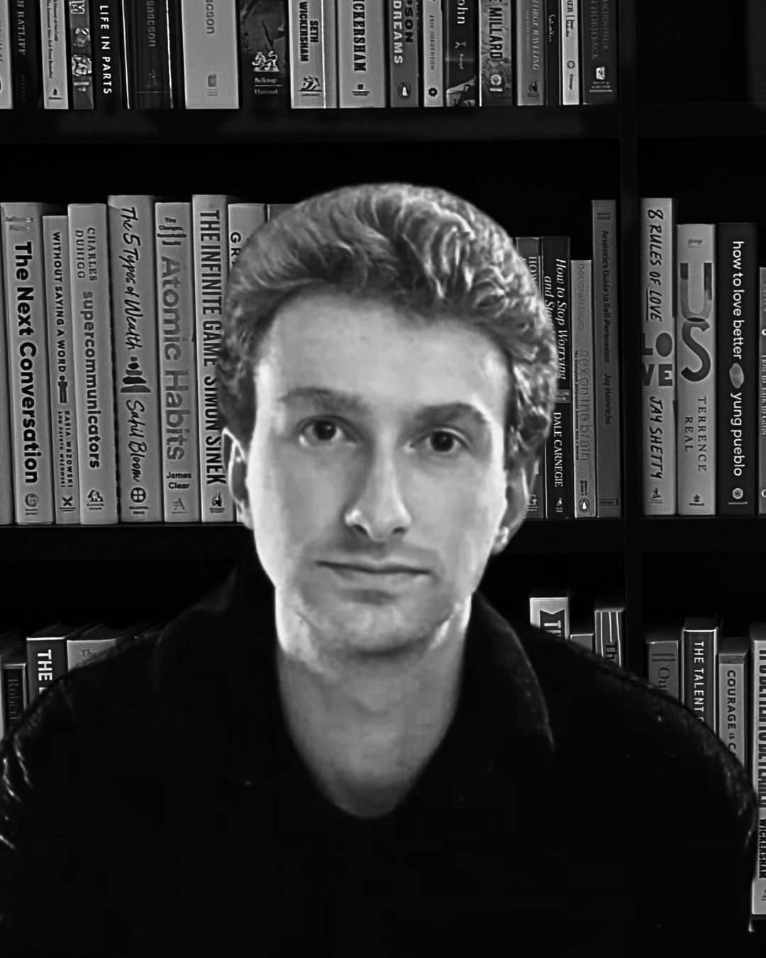 Black and white portrait of a person with short, wavy hair in front of a bookshelf filled with books.