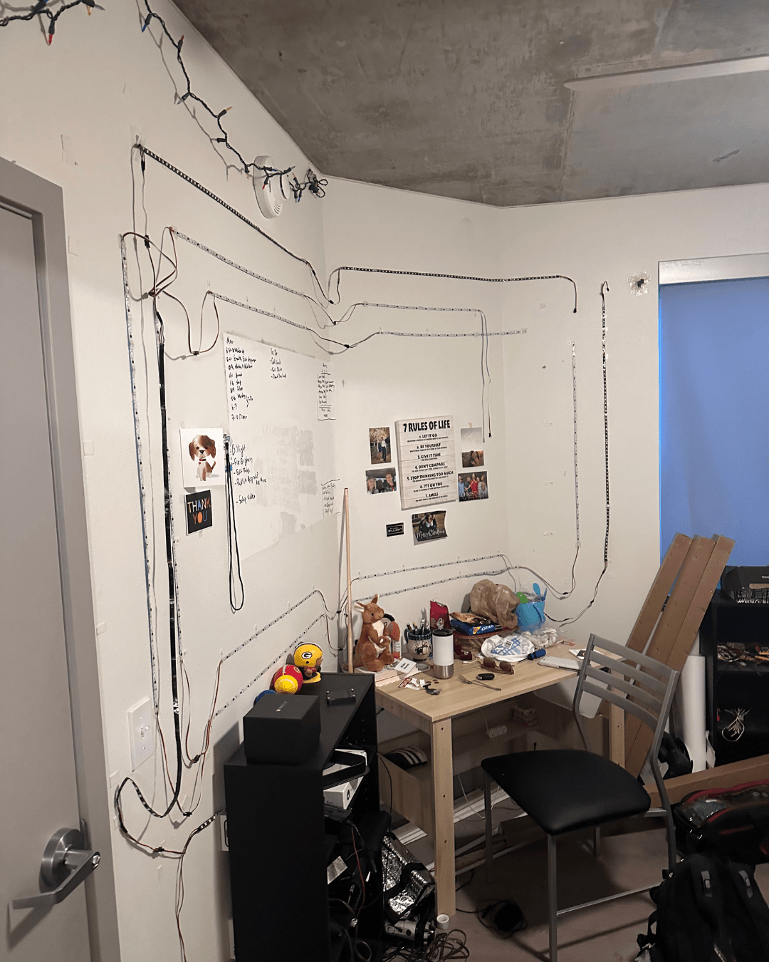 Corner of a room with a desk, chair, and wall decorated with string lights and drawings, window with blue curtain.