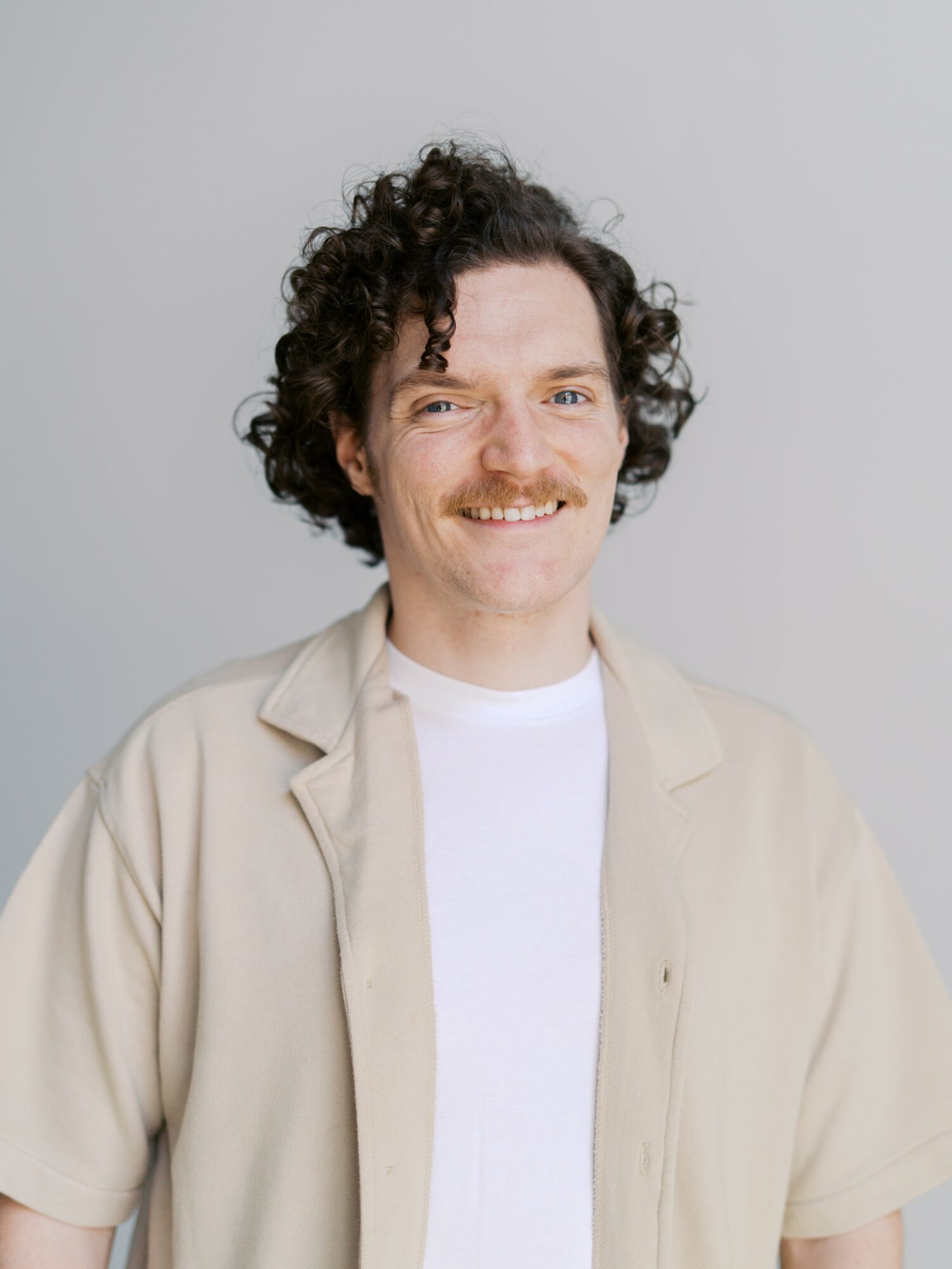Smiling man with curly hair and mustache wearing a beige jacket and white shirt against a plain background.