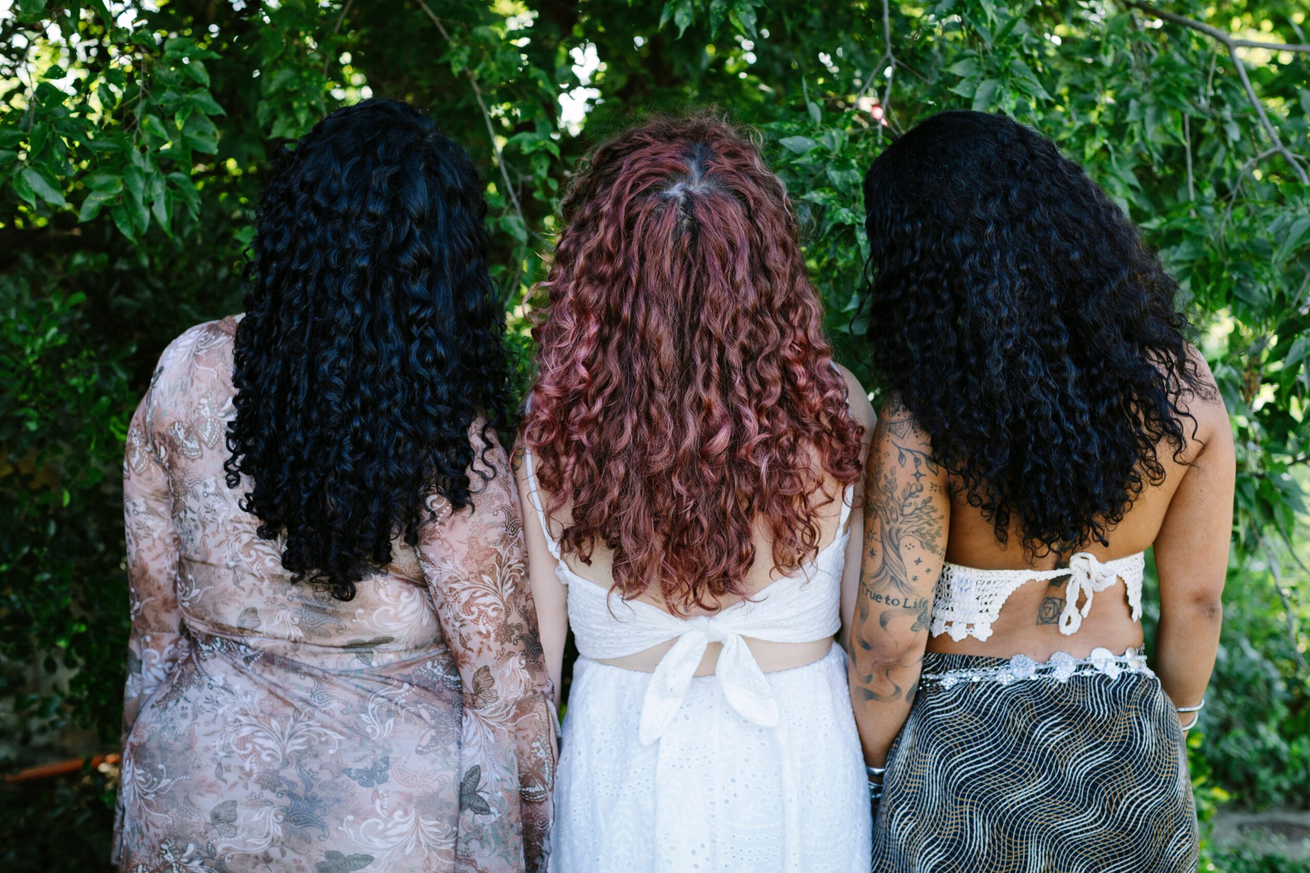Three women with curly hair standing outdoors with green foliage in the background.