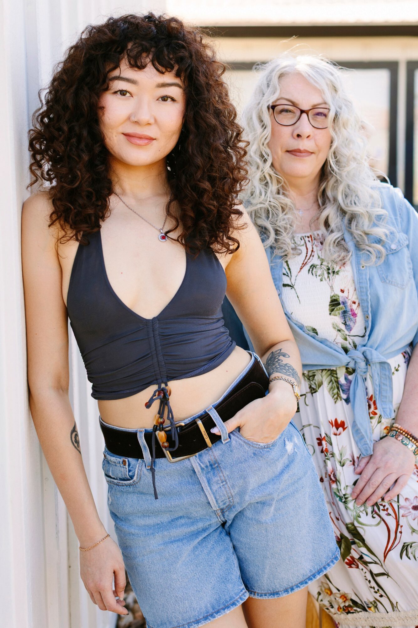 Two women standing outdoors, one with curly dark hair and the other with long gray hair and glasses, smiling.