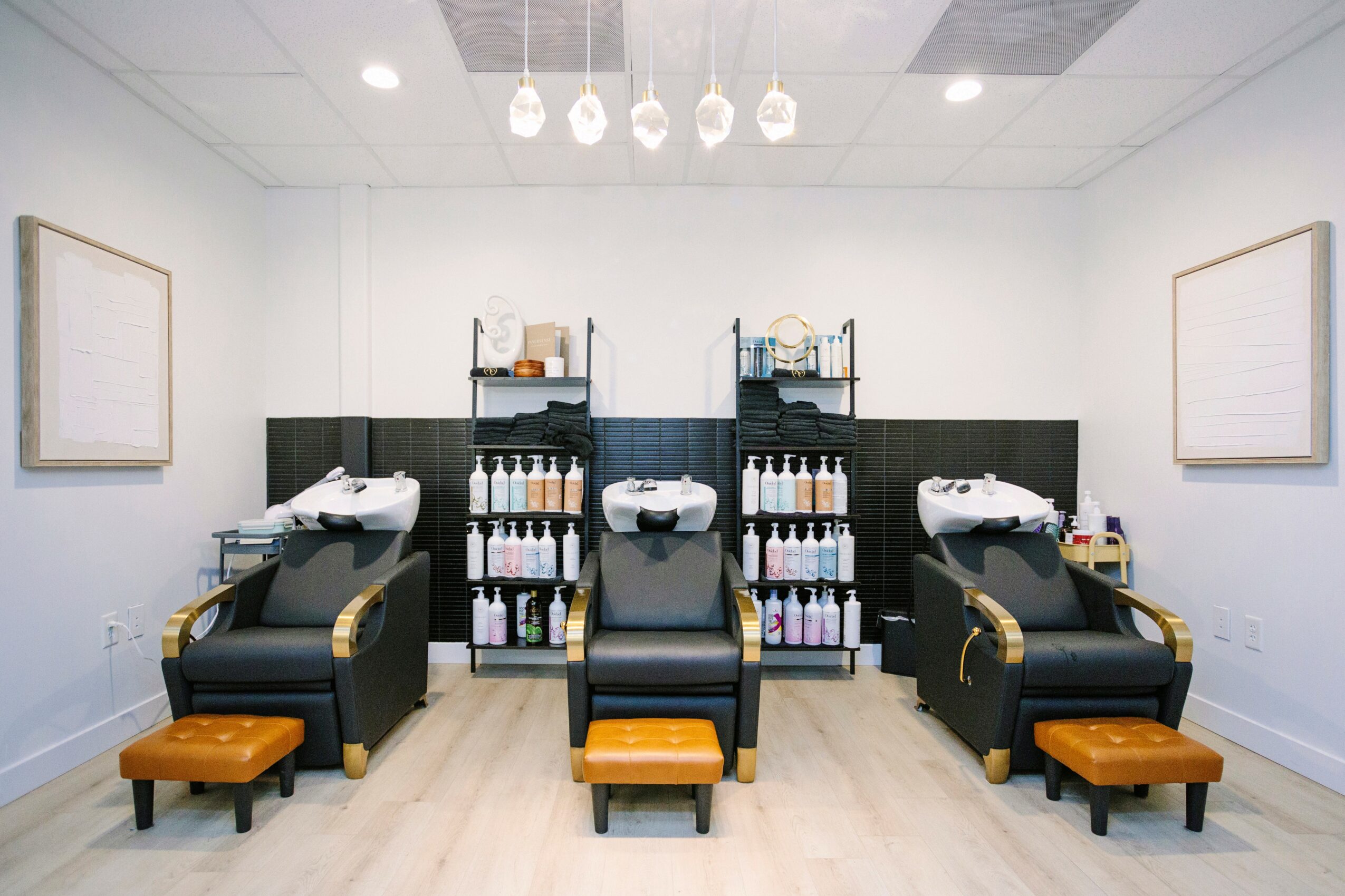 Three salon chairs with footrests and shelves holding bottles, in a bright, white room with framed artwork.
