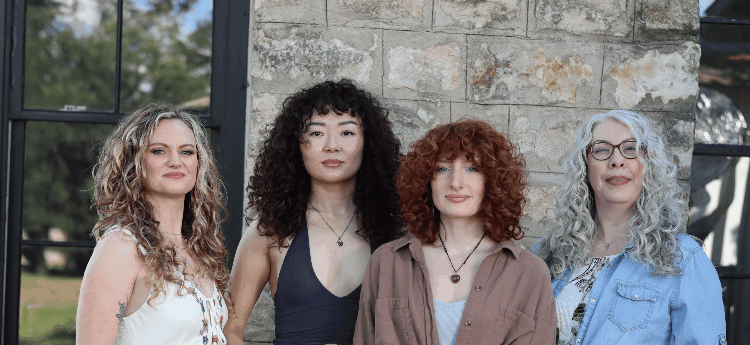 Four women standing outdoors in front of a stone wall and window, smiling and looking at the camera.
