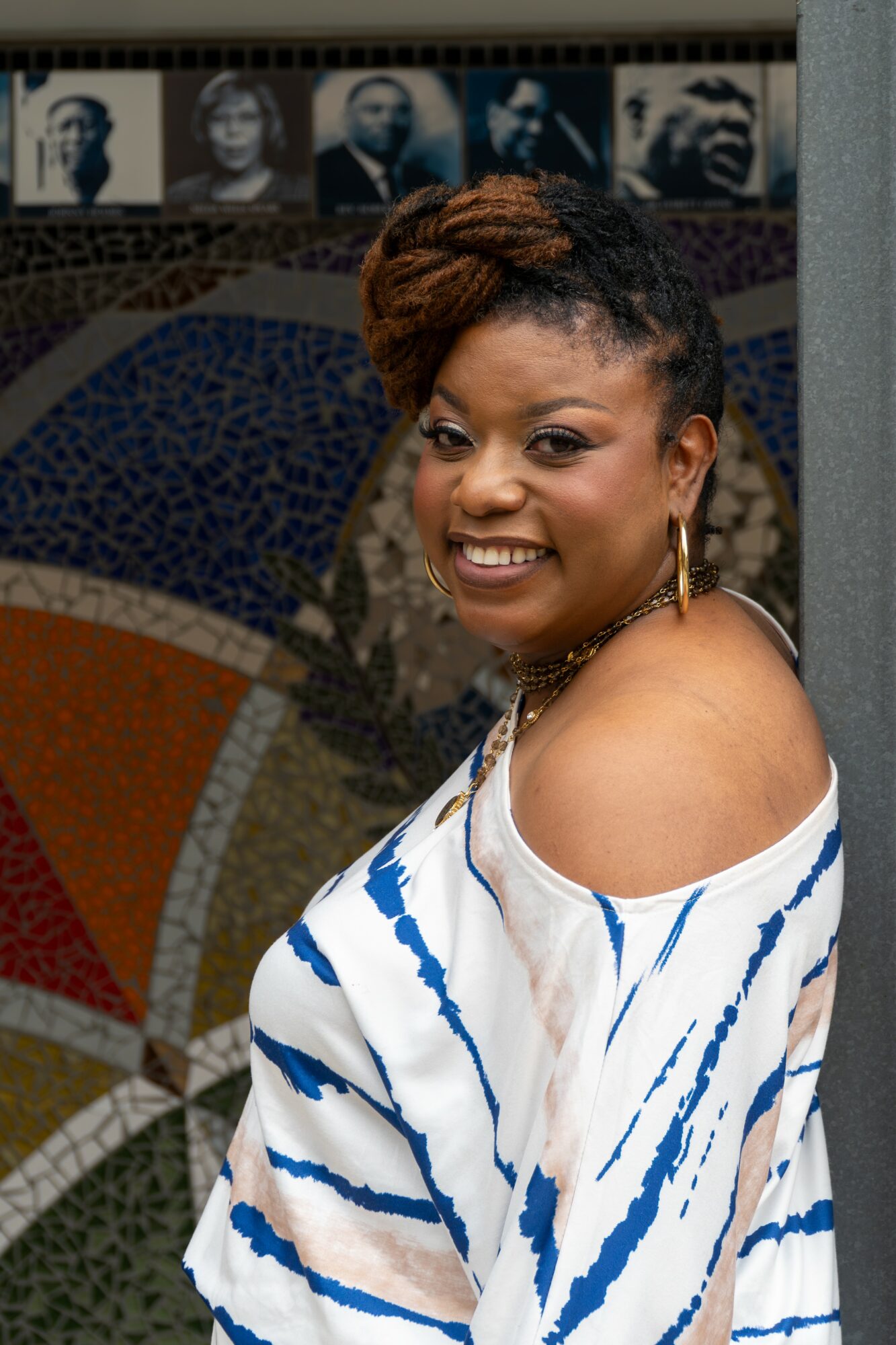 Smiling woman with short hair and earrings, wearing a white and blue striped off-shoulder top, standing indoors.