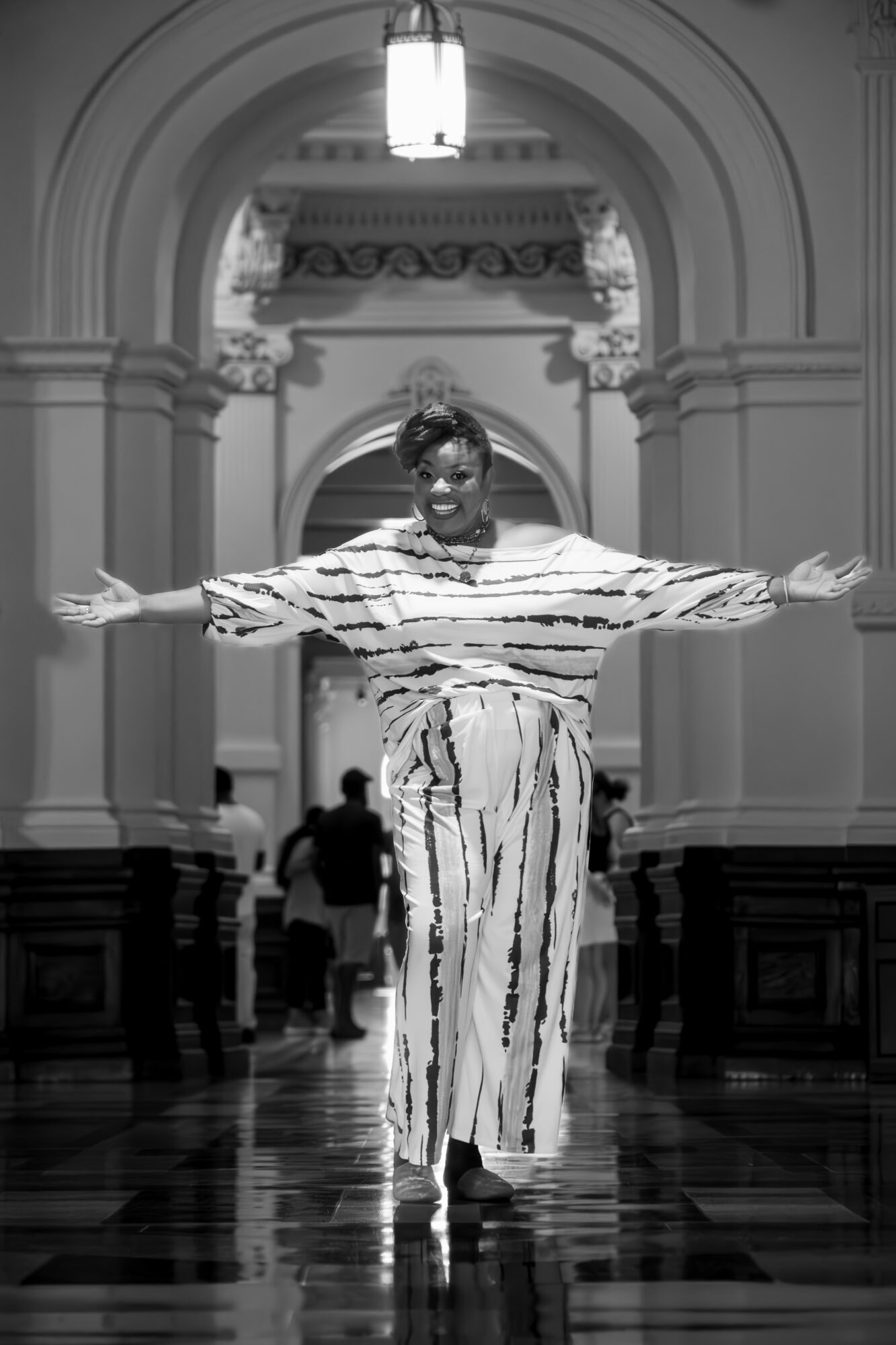 Person standing with arms outstretched in a grand hallway with arches and ornate details, wearing striped clothing.