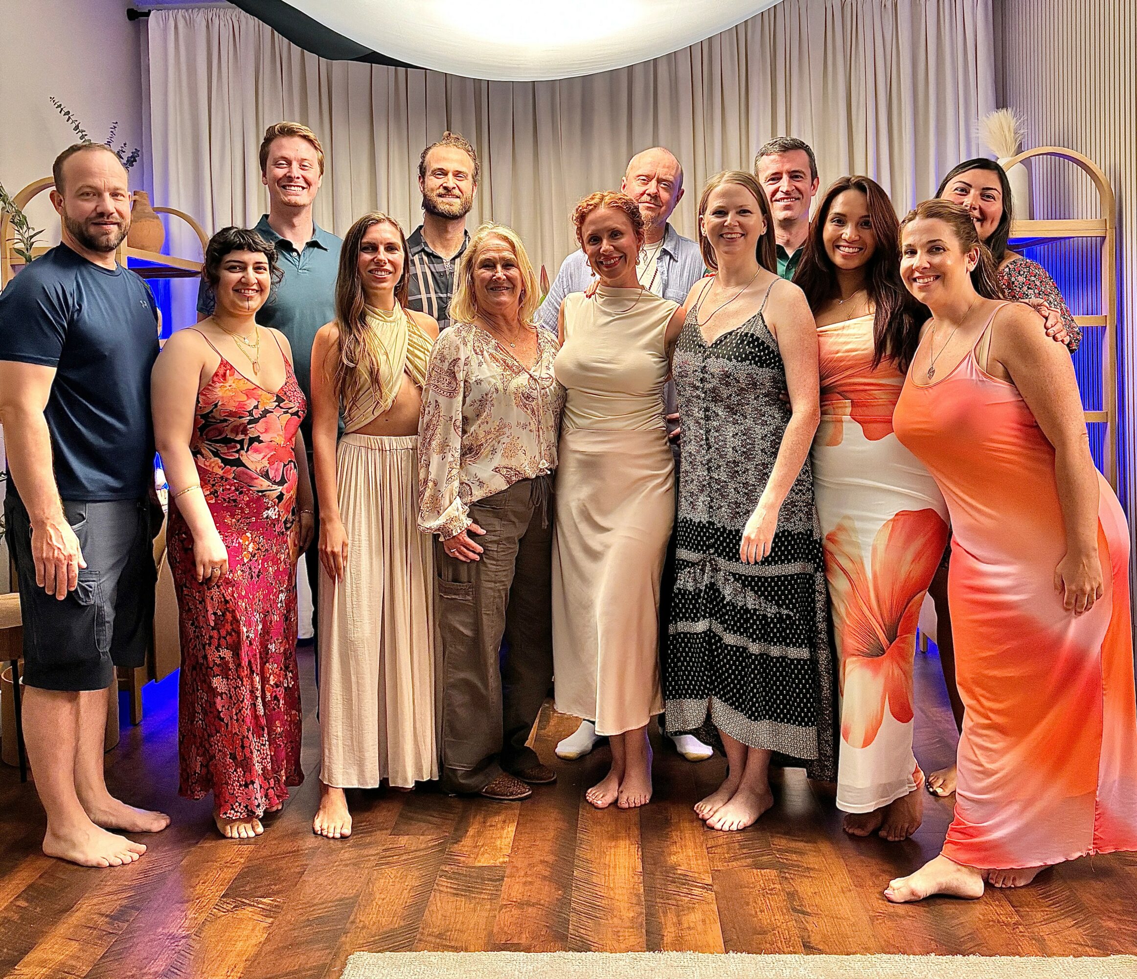 Group of 14 people standing indoors, smiling, with curtains and a wooden floor background.