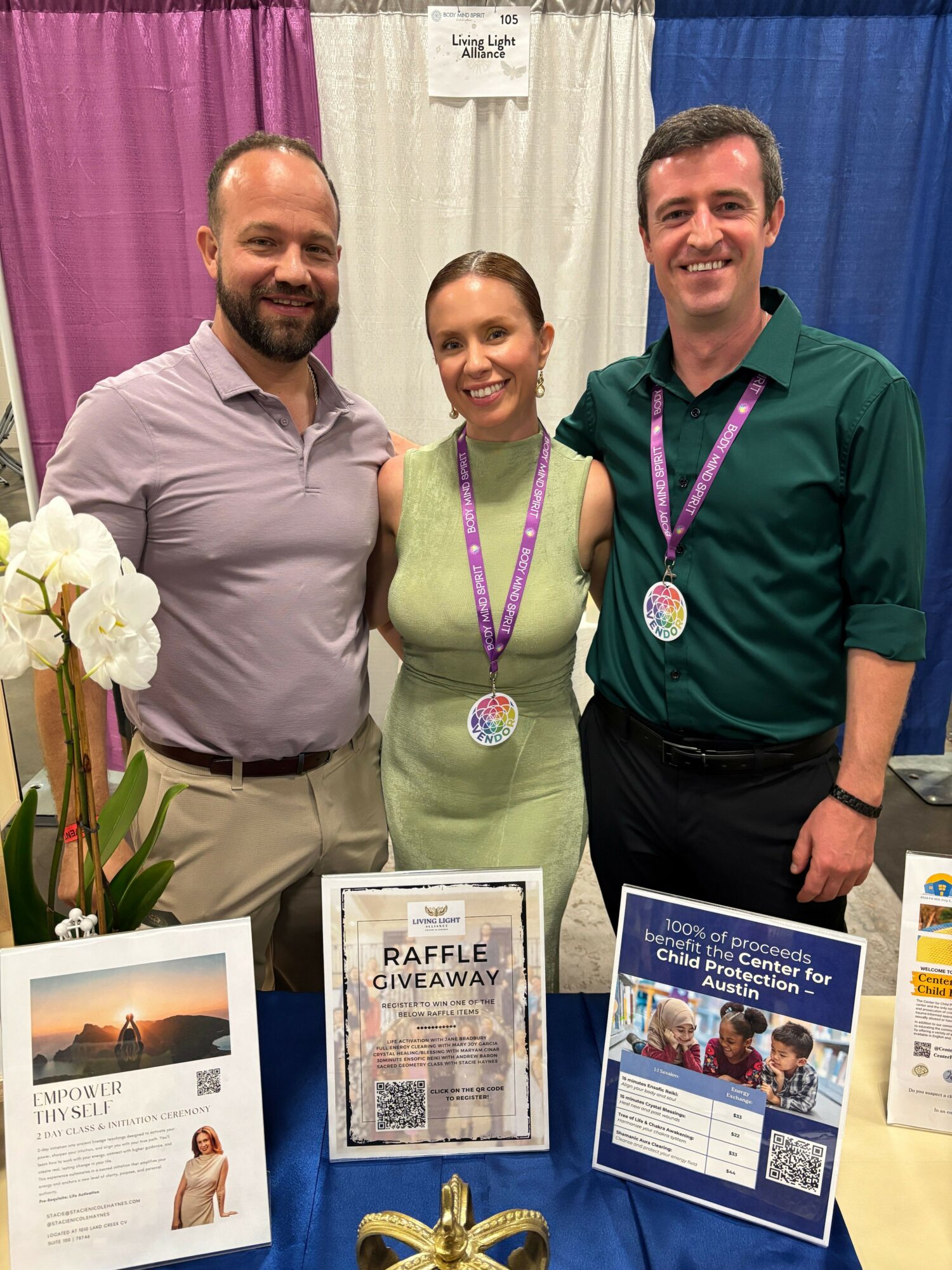Three people standing behind a table with informational materials, flowers, and a purple curtain backdrop.