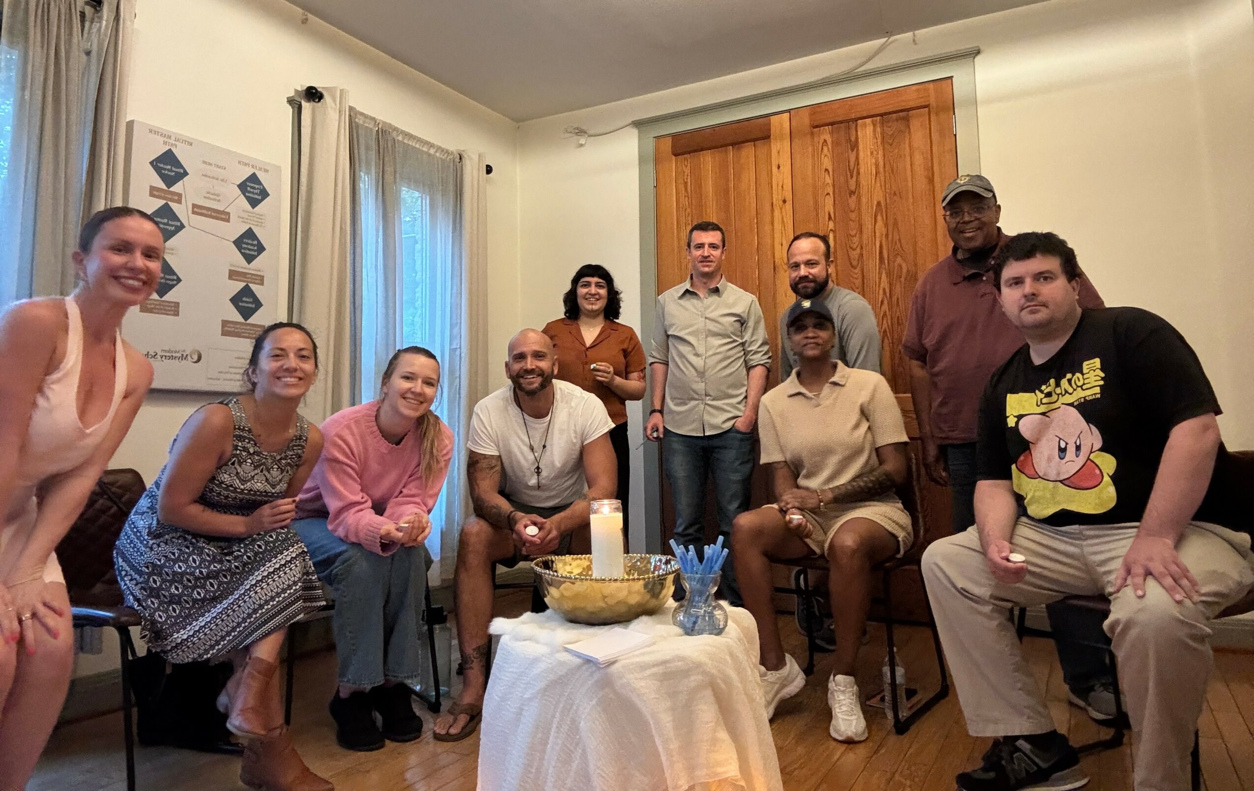 Group of ten people gathered around a table with a cake, smiling indoors with curtains and wooden door behind.