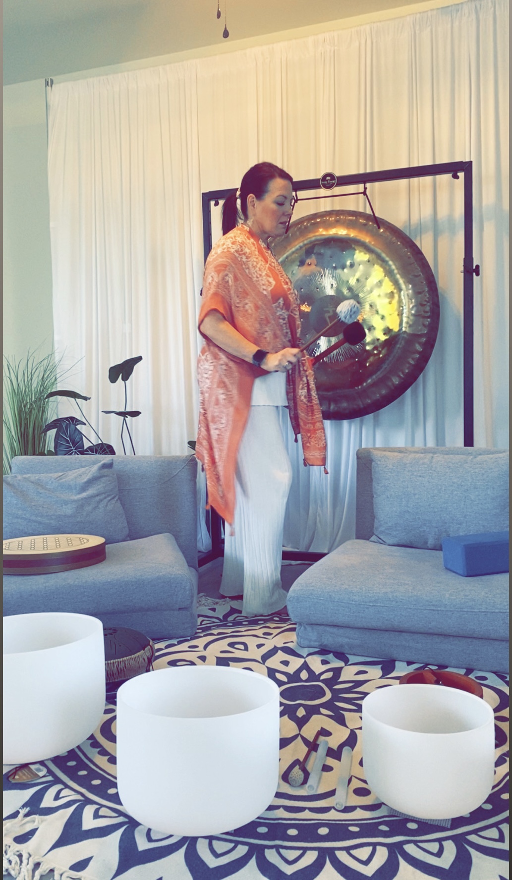 Woman in traditional clothing standing near a large gong in a living room with curtains and furniture.