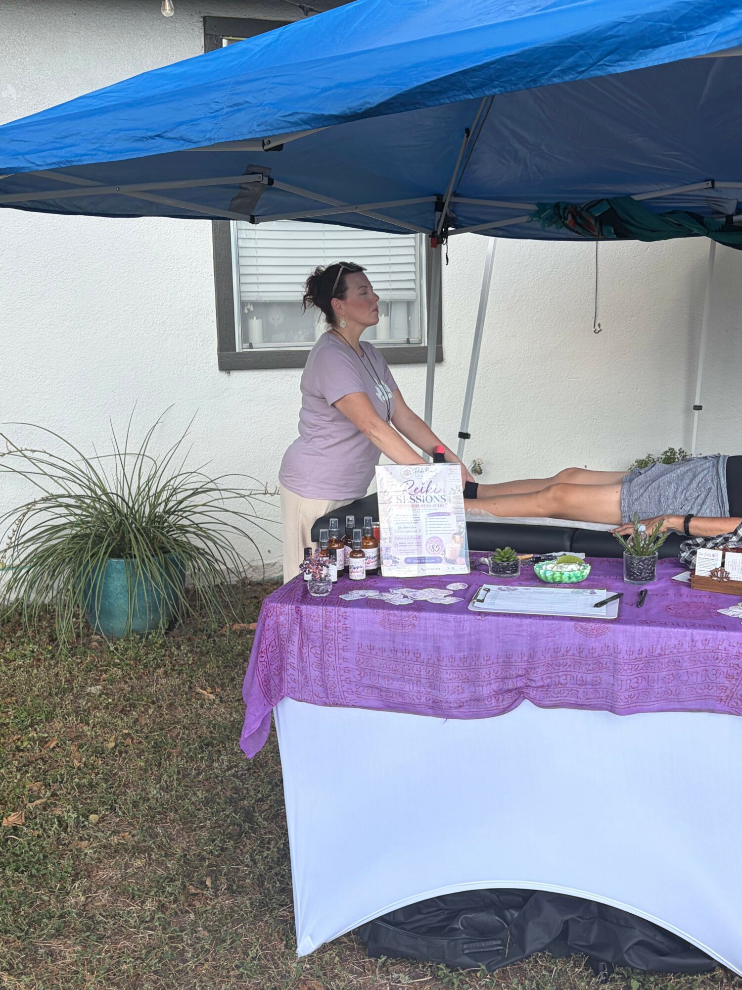 Person standing at a table with a purple cloth, outdoors, with a potted plant nearby.