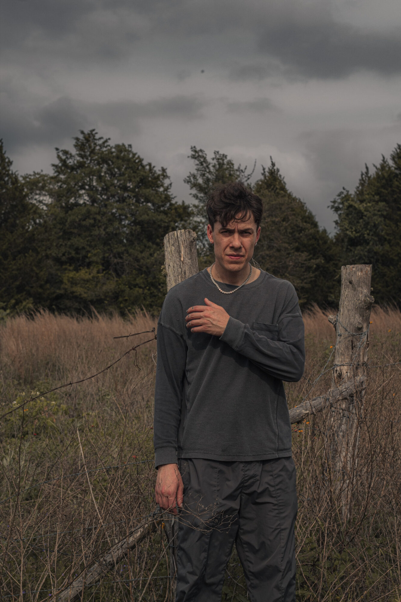 Young man standing outdoors in a field with trees and a cloudy sky, wearing a dark long-sleeve shirt and pants.