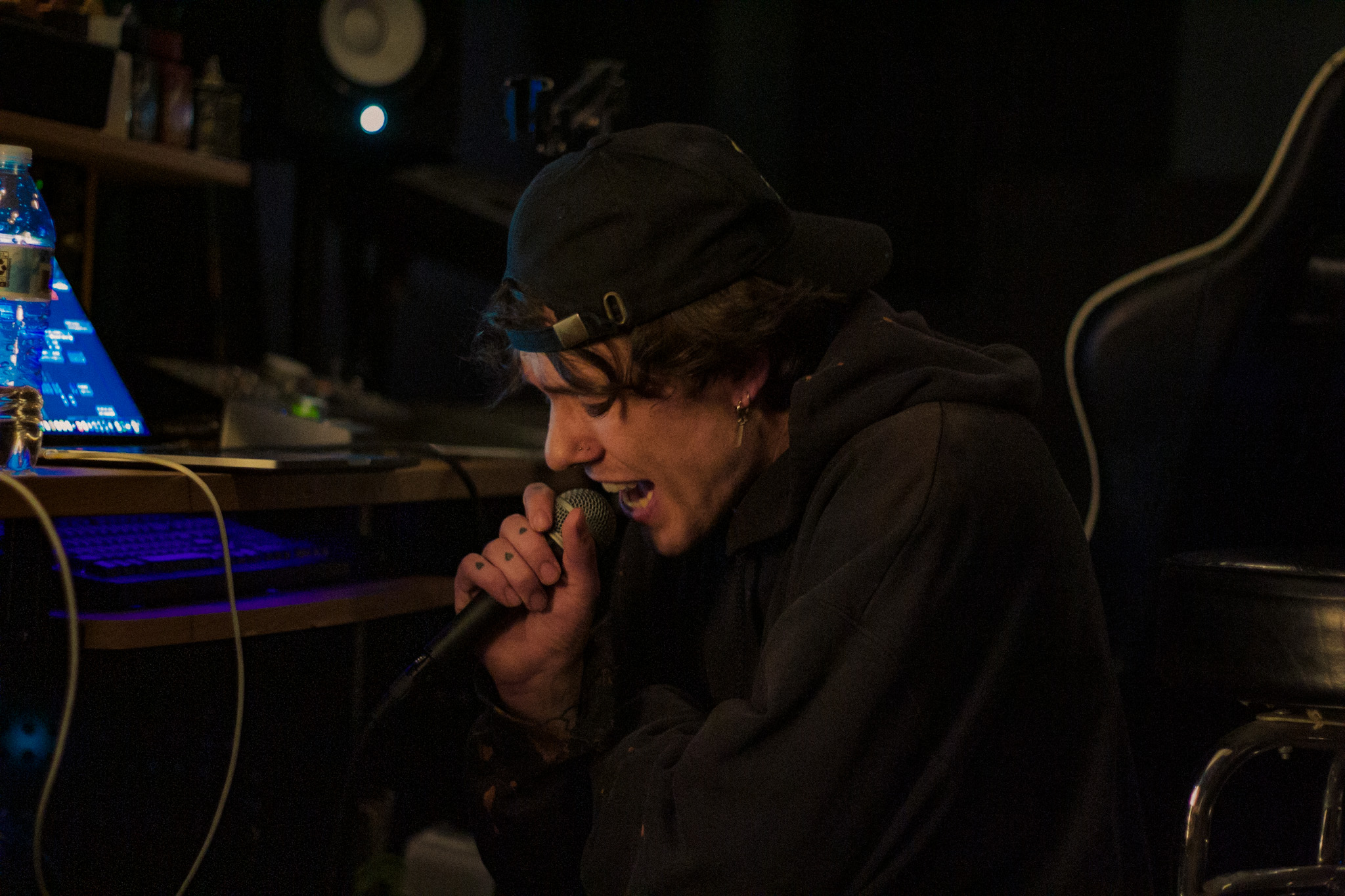 Person singing into a microphone in a dark room with electronic equipment, wearing a black hoodie and cap.
