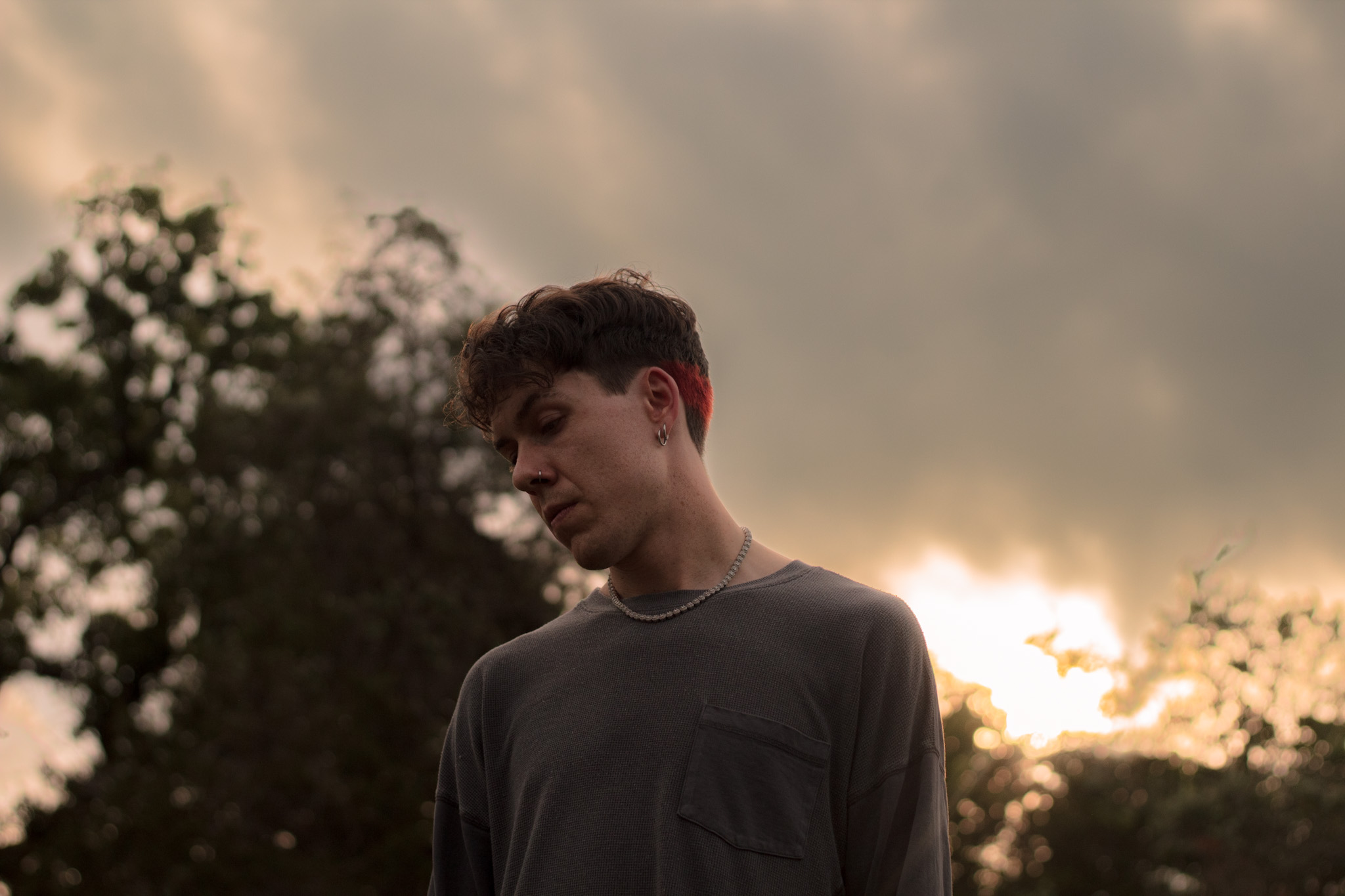 Young man with dark hair wearing a dark shirt outdoors, cloudy sky in background.