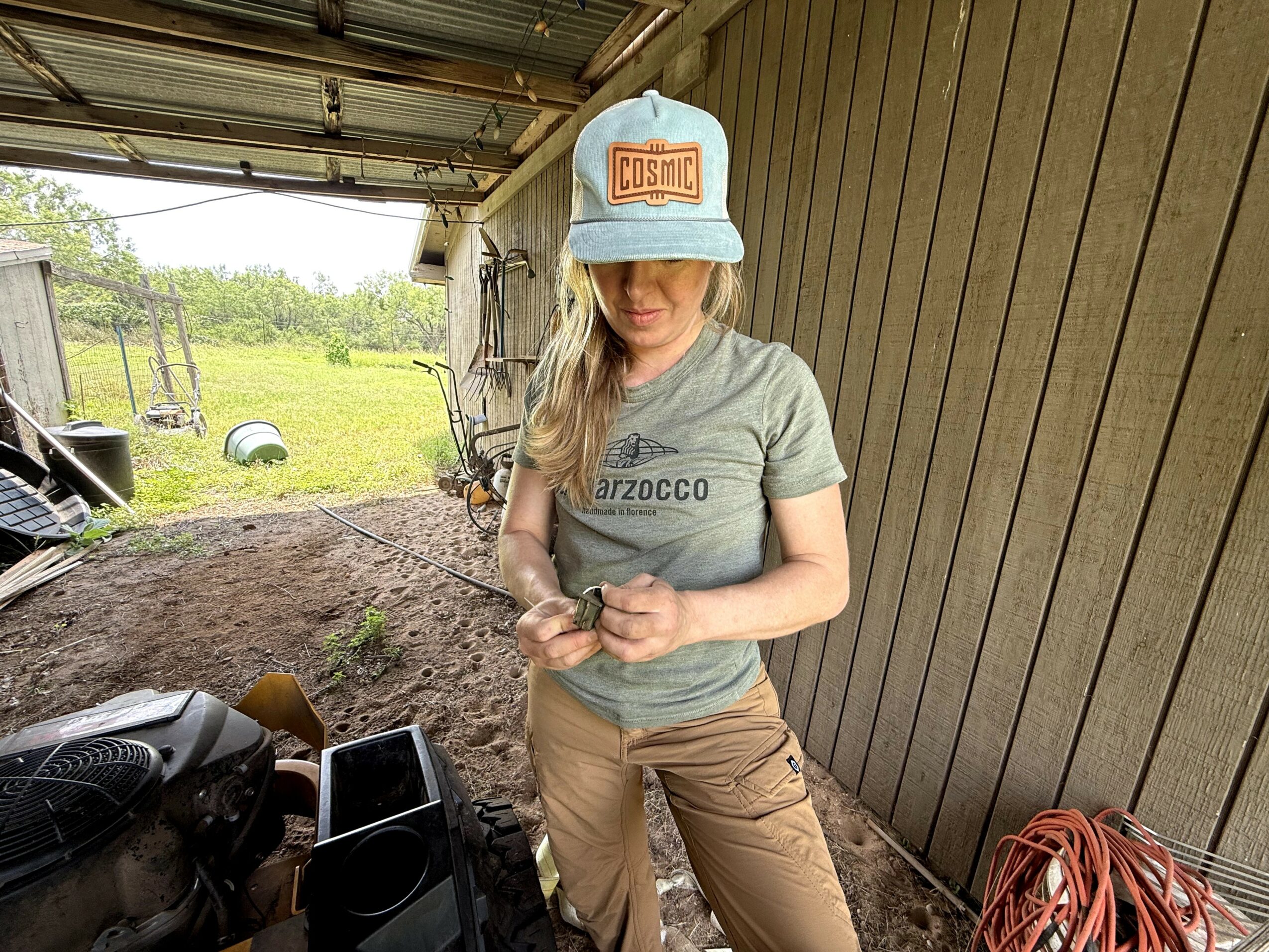 Woman wearing a cap and casual clothes standing in a garage with tools and a grassy outdoor area visible behind her.