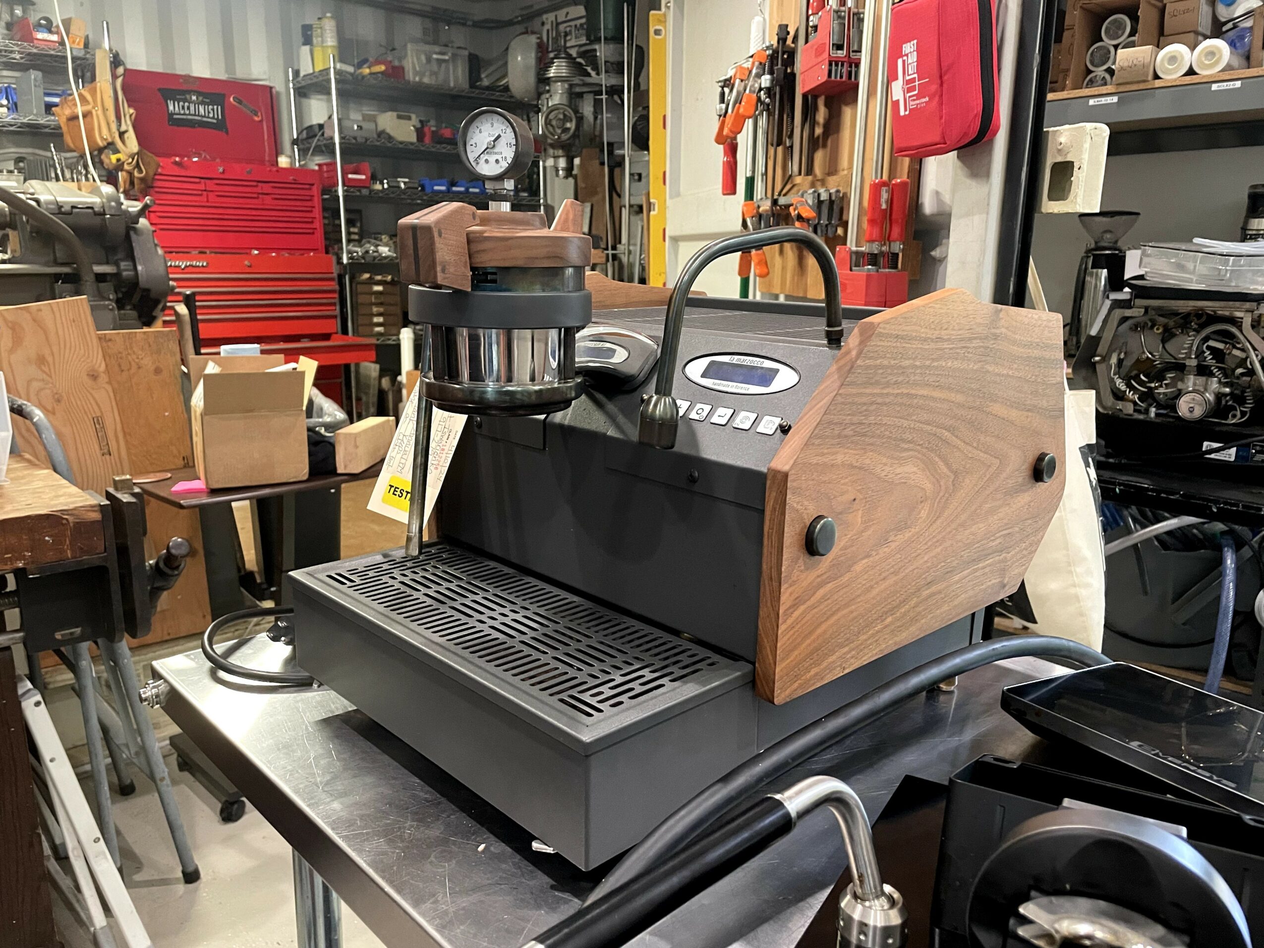 Coffee machine with wooden side panels and a metal drip tray, in a workshop setting with tools and equipment in the background.