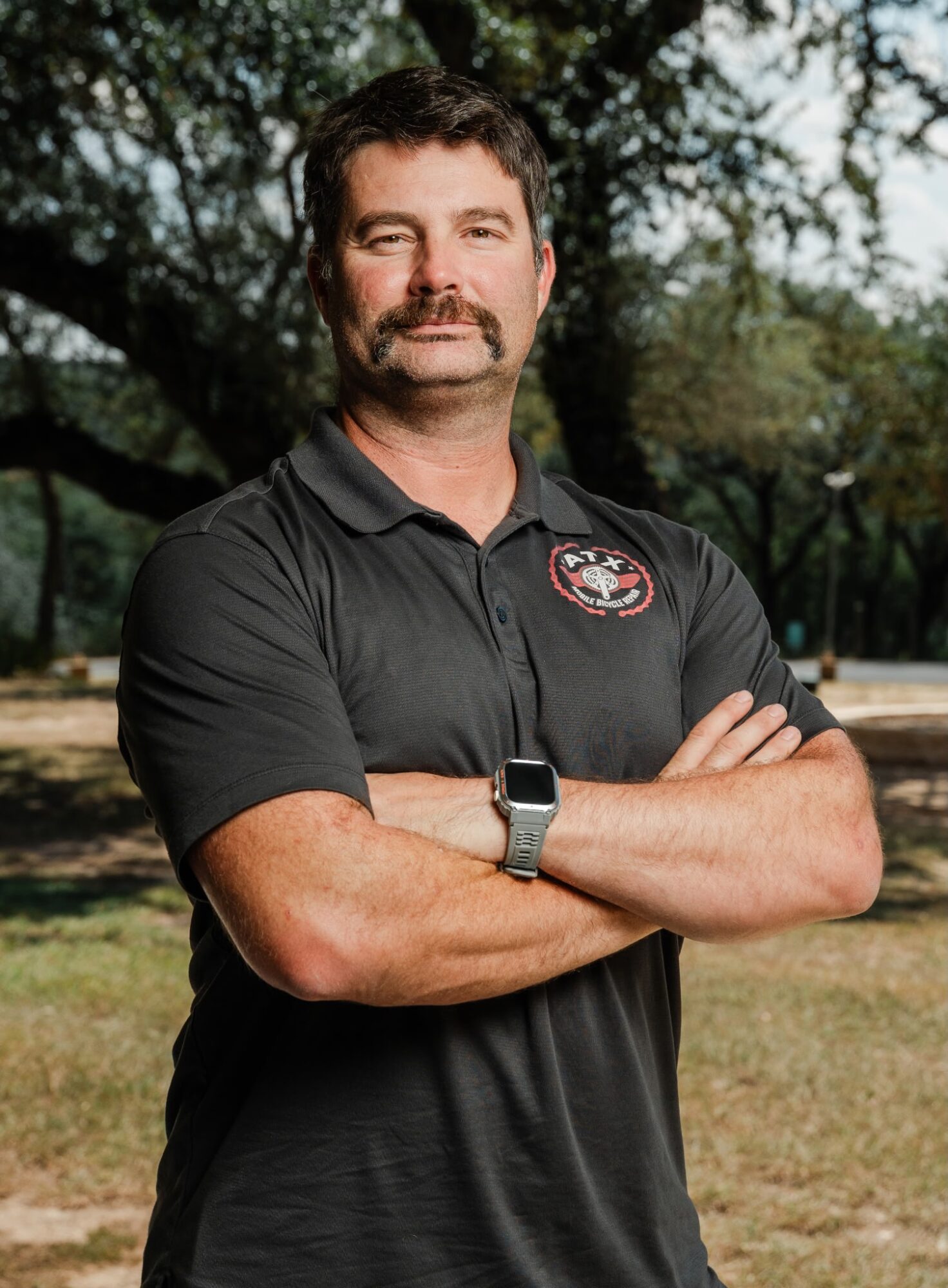 Man with crossed arms, wearing a black polo shirt with a logo, outdoors with trees and grass in background.