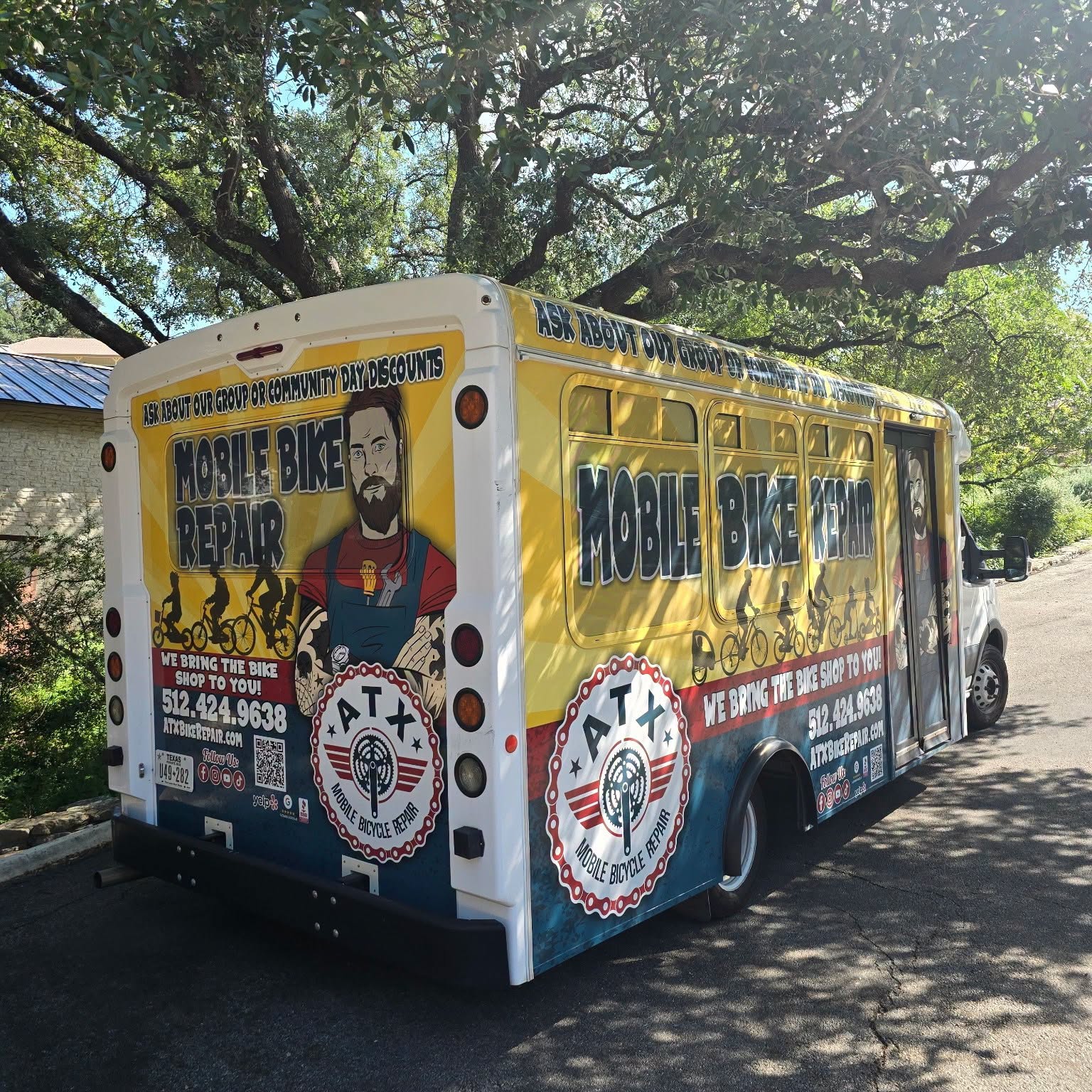 Mobile bike repair bus parked outdoors with trees in background, displaying service information and logos on yellow and white vehicle.