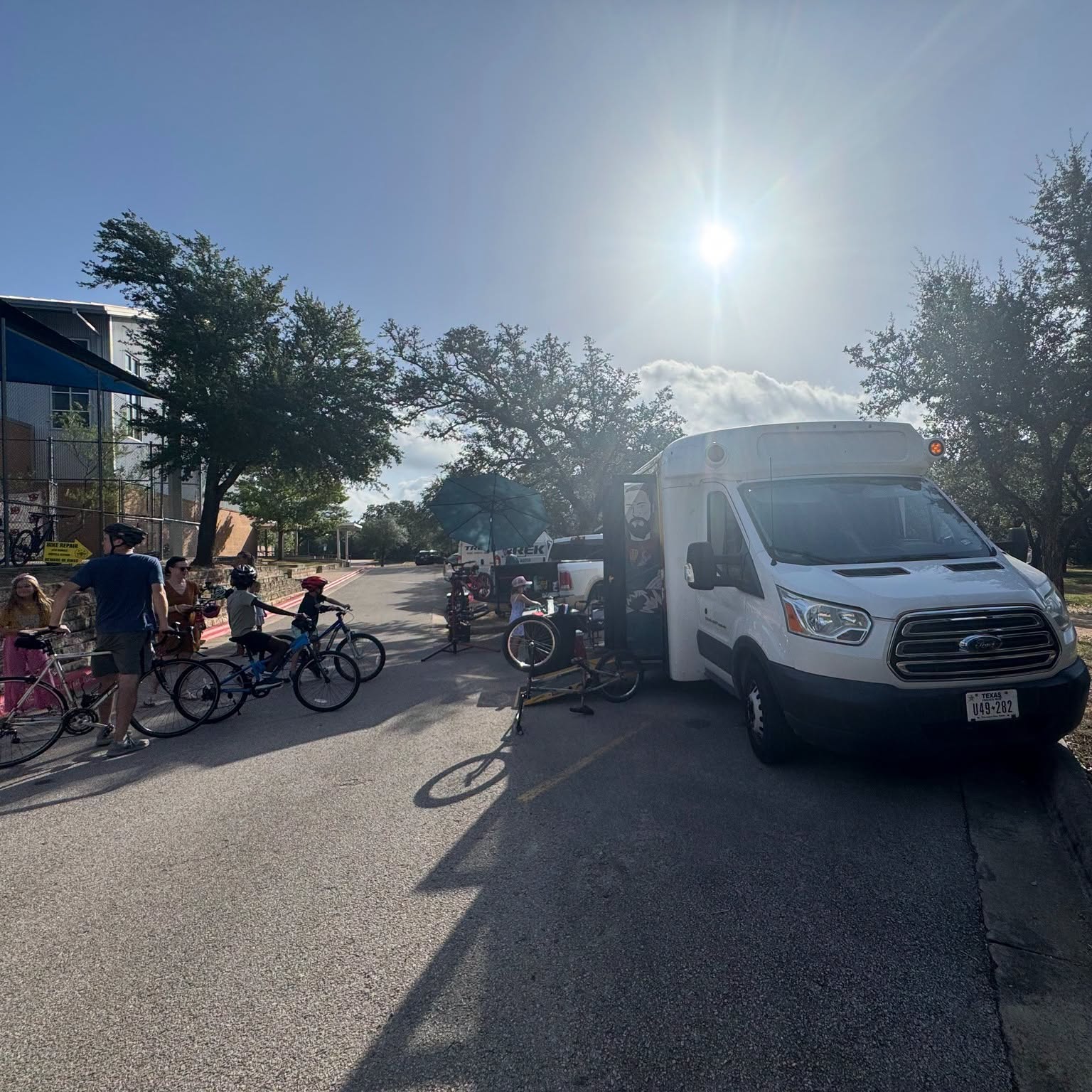 People gather around a white van parked on the street with trees and a building in the background, sunlight shining overhead.