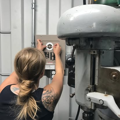 Person with a tattoo on their arm adjusting a control panel on a wall, industrial equipment nearby.