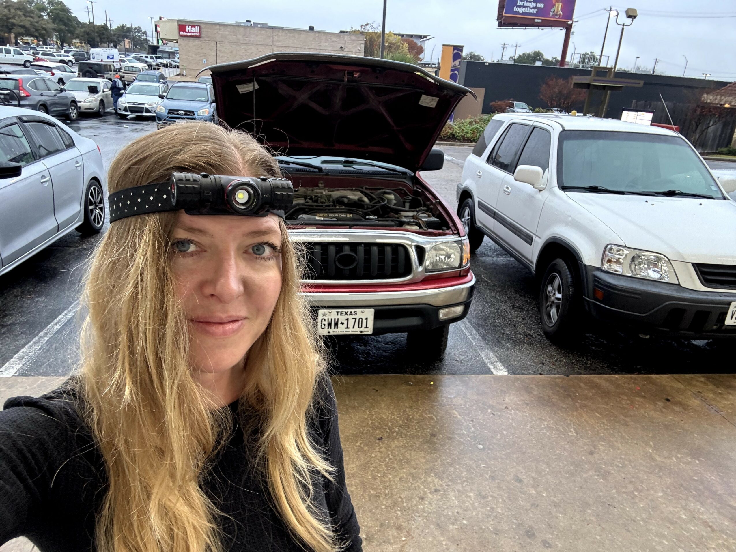 Woman with long hair wearing a headlamp taking a selfie in a parking lot with cars and a car with open hood behind her.