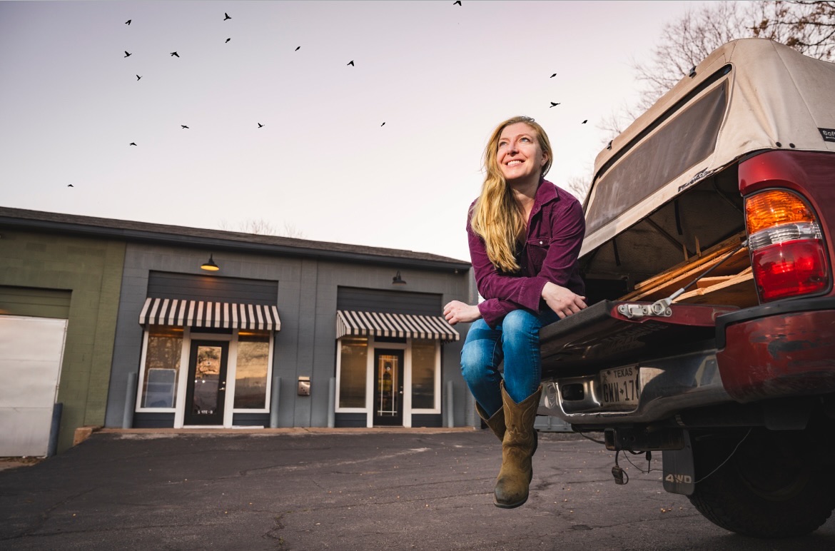 Young woman sitting on the open tailgate of a pickup truck, smiling, in front of a building with striped awnings.