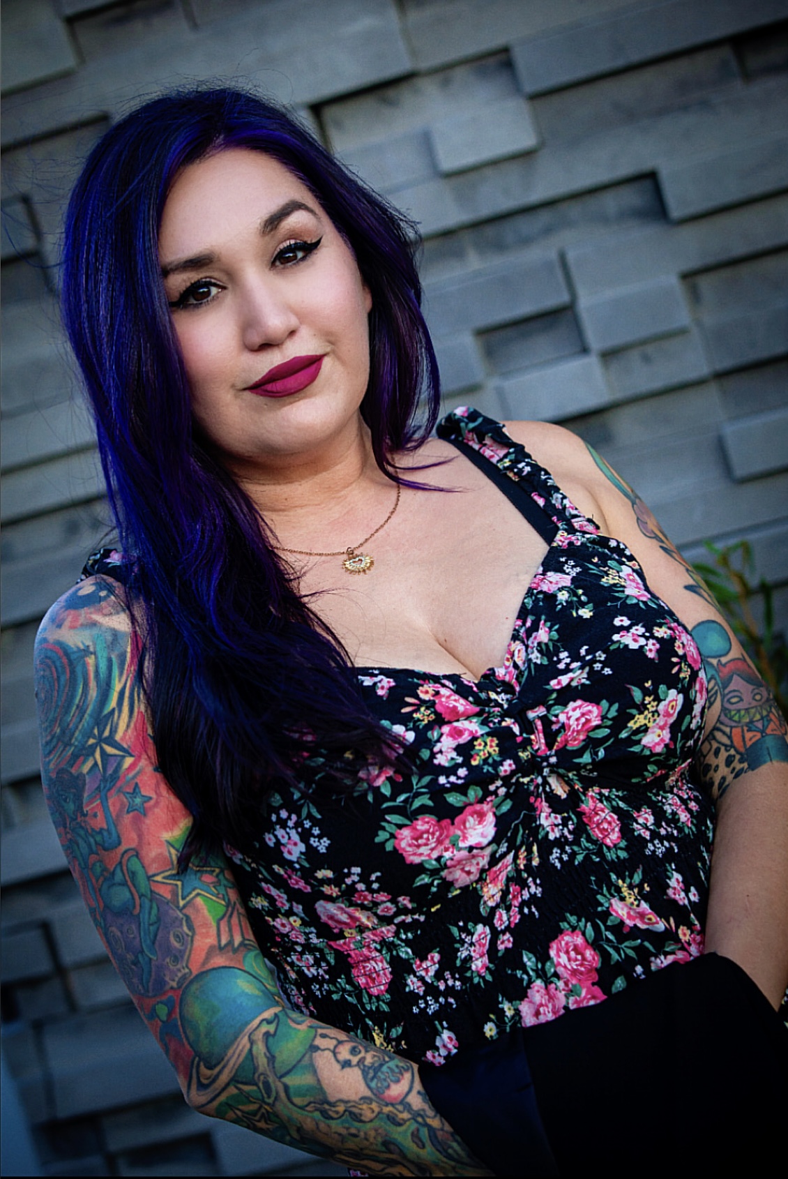 Woman with long dark blue hair and colorful tattoos on her arms, wearing a floral top, standing outdoors against a stone wall.