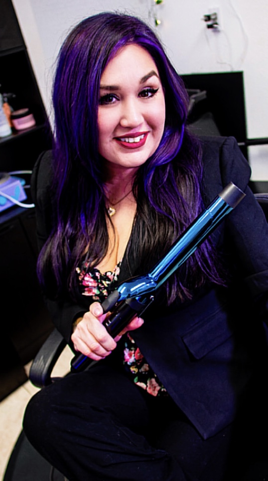 Woman with purple hair smiling, holding a curling iron, sitting in a room with shelves and a computer.