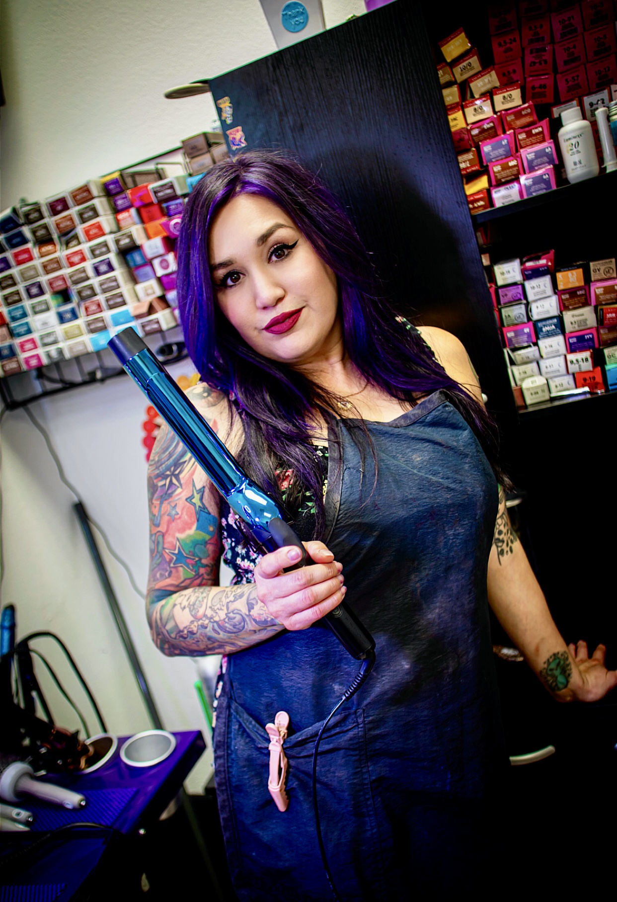Woman with purple hair and tattoos holding a blue hairdryer in a salon, shelves with hair products behind her.