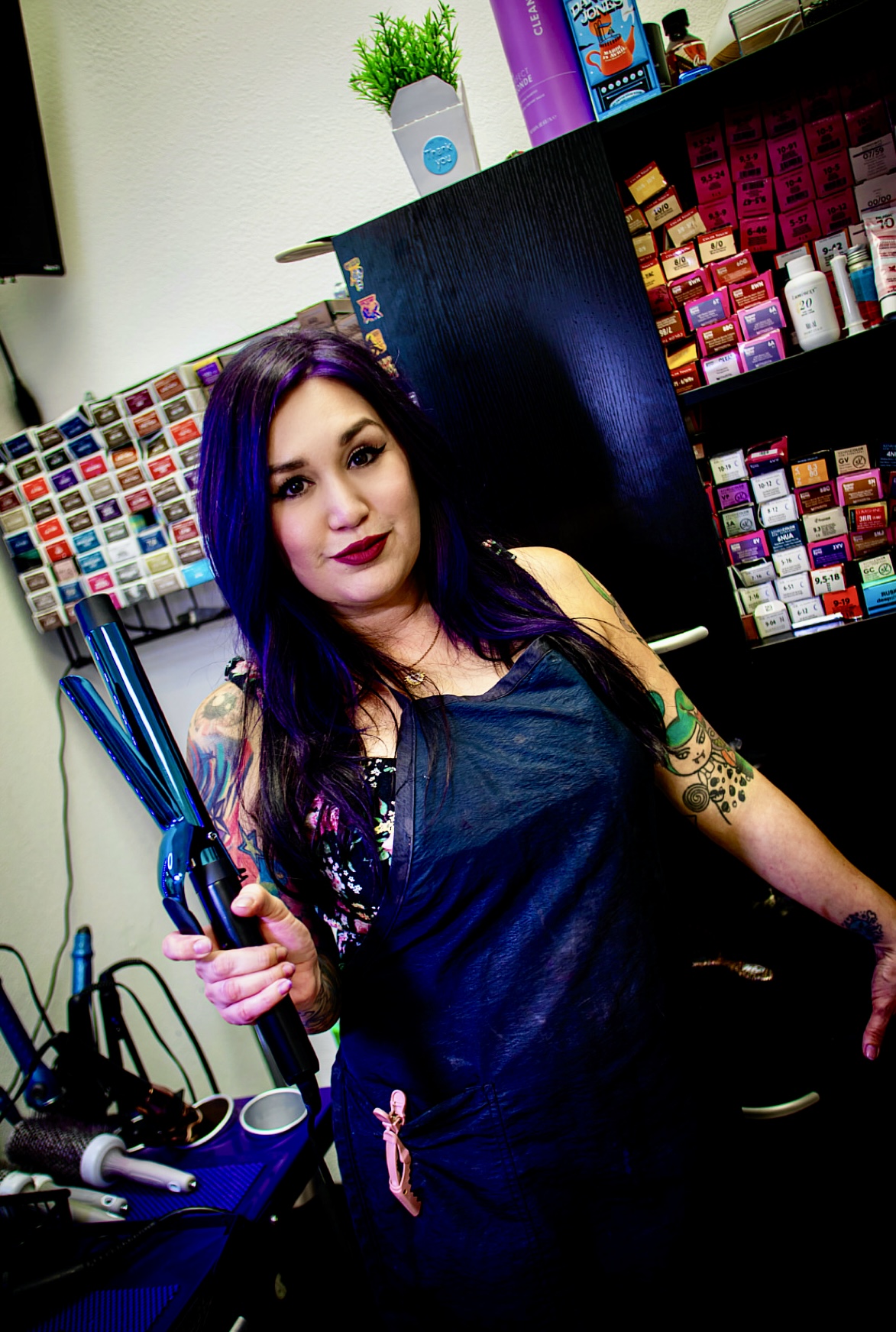 Woman with dark hair and tattoos holding hair styling tools in a salon, colorful shelves in background.