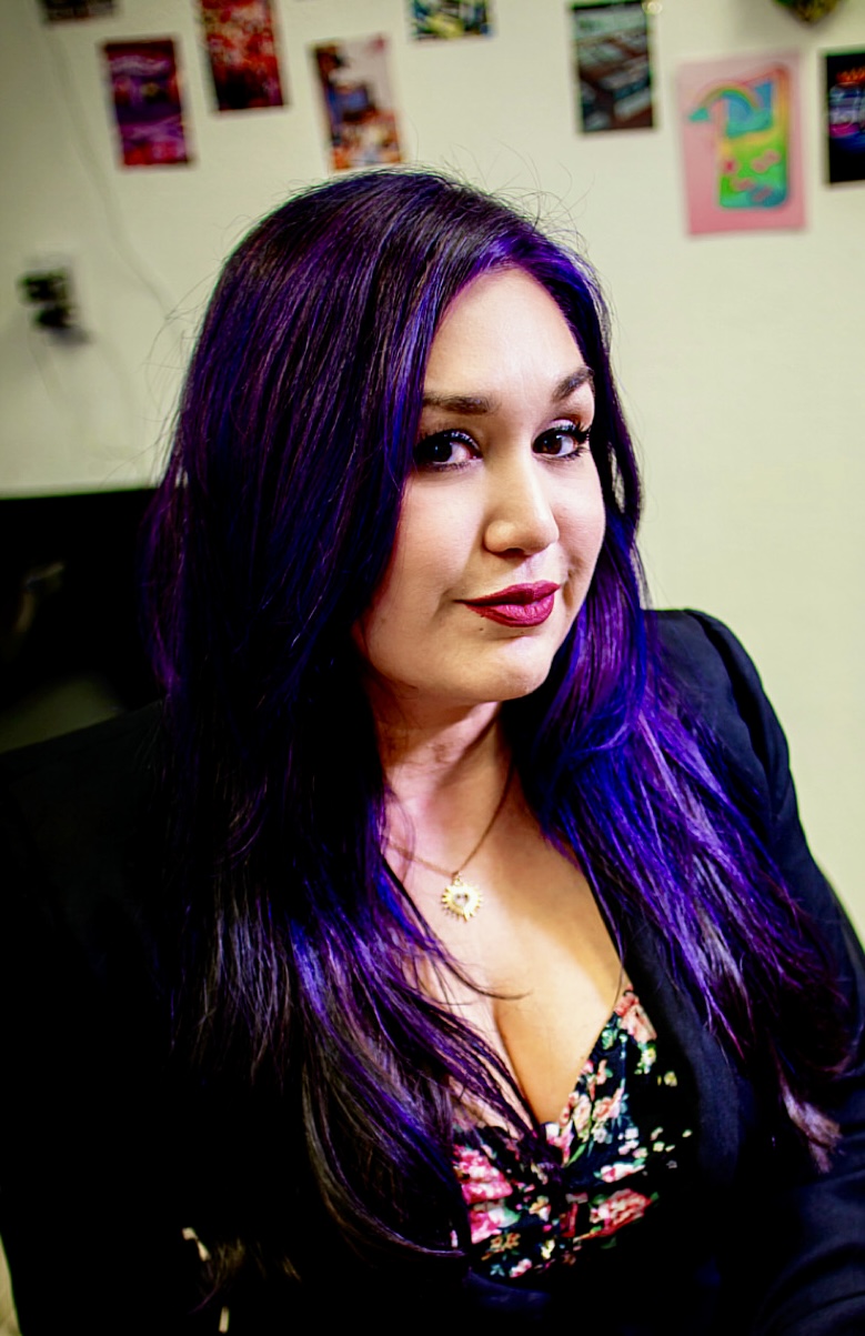 Woman with long purple and black hair, wearing a floral top and black jacket, smiling in an indoor setting.
