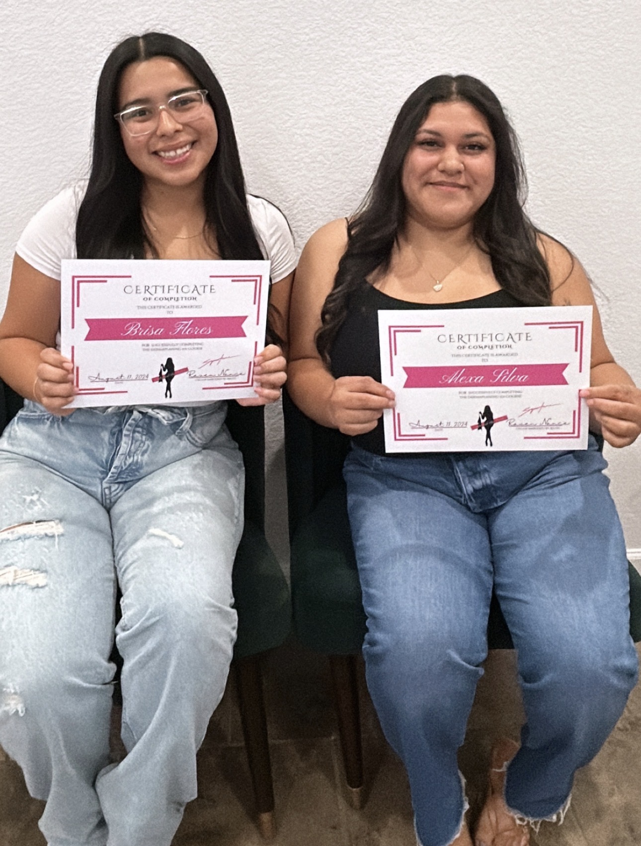 Two women sitting on chairs holding certificates, smiling, against a plain wall, wearing casual clothing.