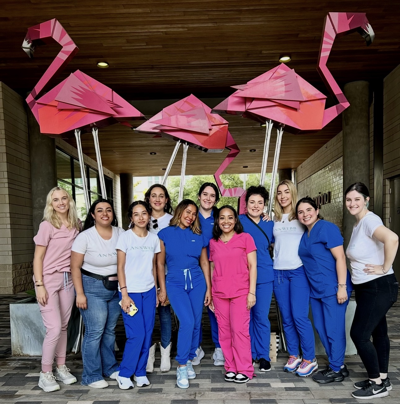 Group of women standing indoors with pink flamingo decorations hanging from the ceiling, smiling, some wearing scrubs.