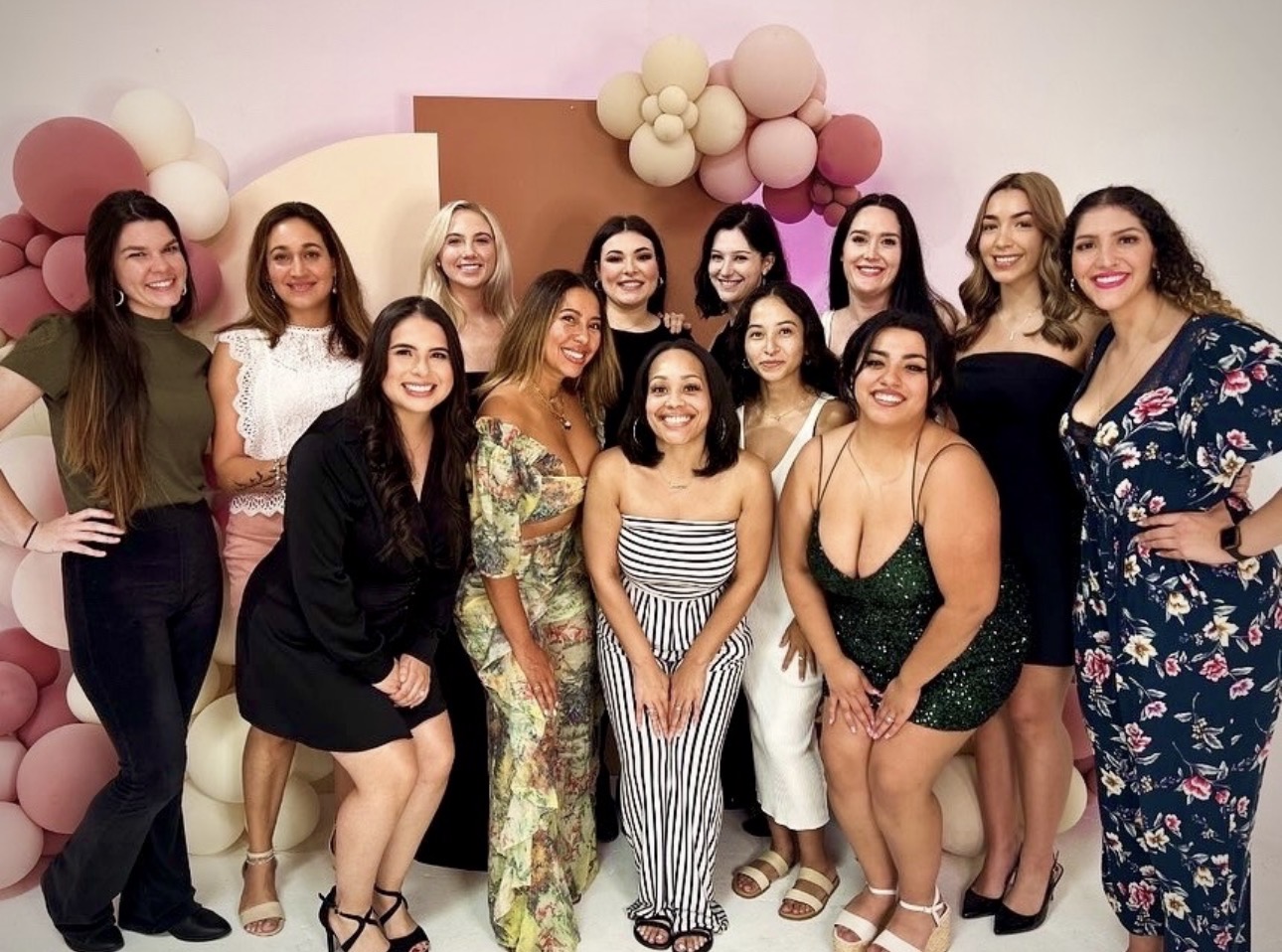 Group of twelve women smiling, standing in front of pink and beige balloon decorations, posing for a photo.