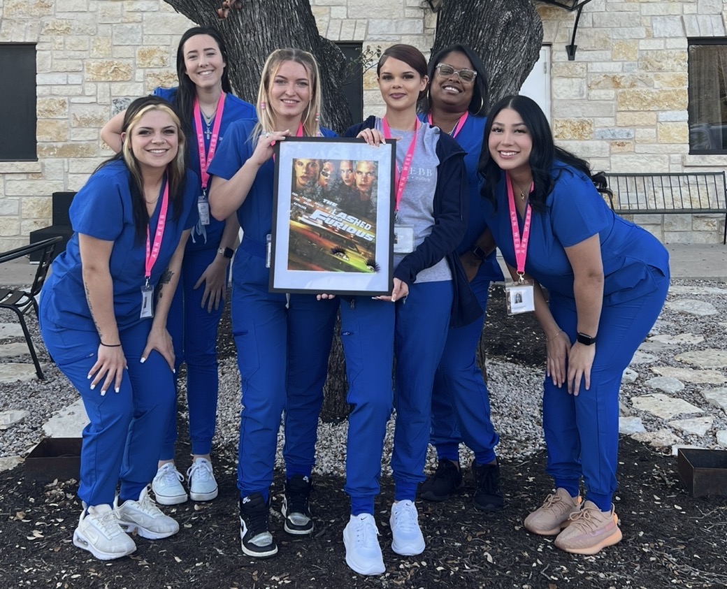 Six healthcare workers in blue scrubs holding a framed photo outdoors, smiling, with trees and a stone building in background.