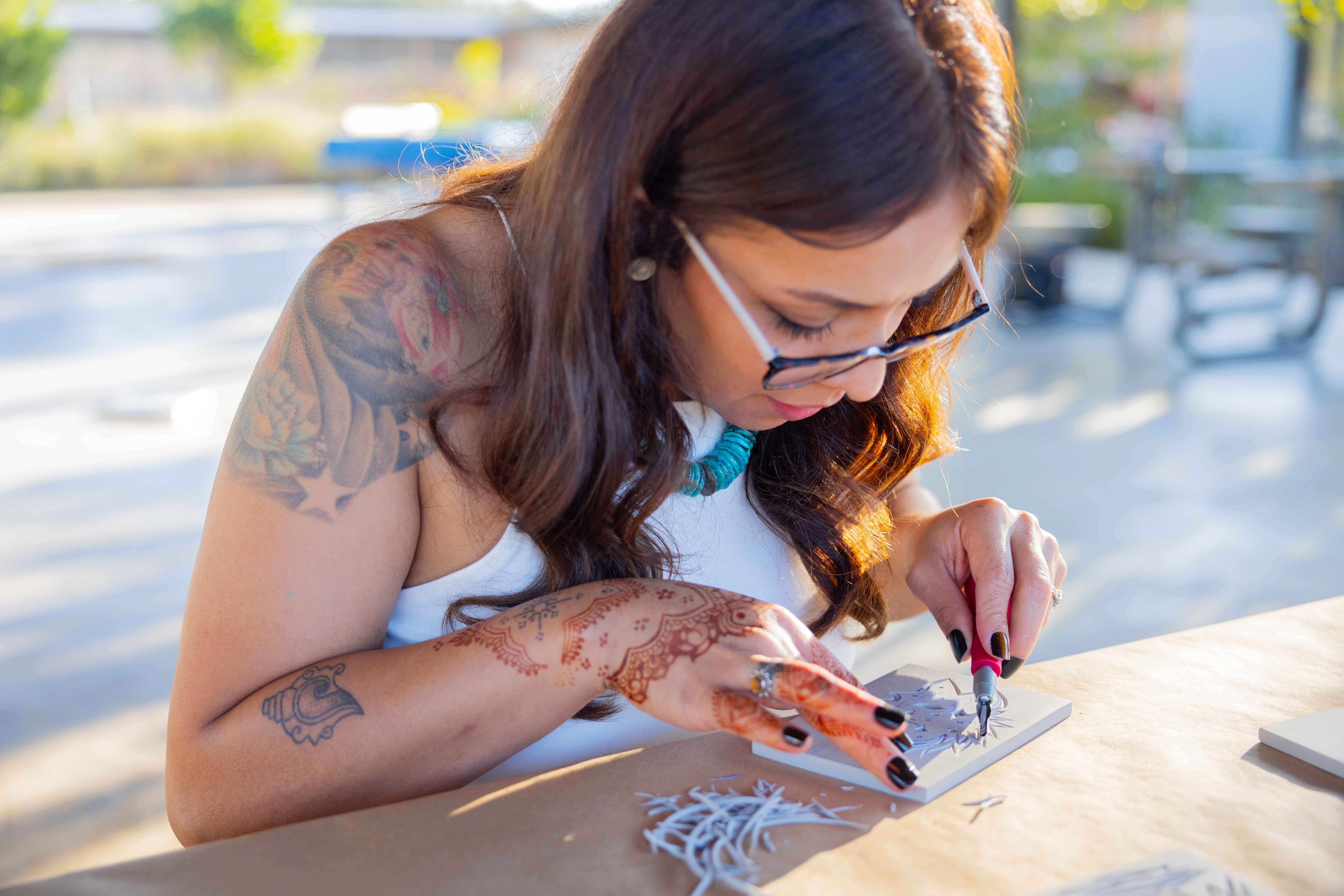 Woman with glasses and tattoos, focused on her hand, working on a craft project outdoors at a table.