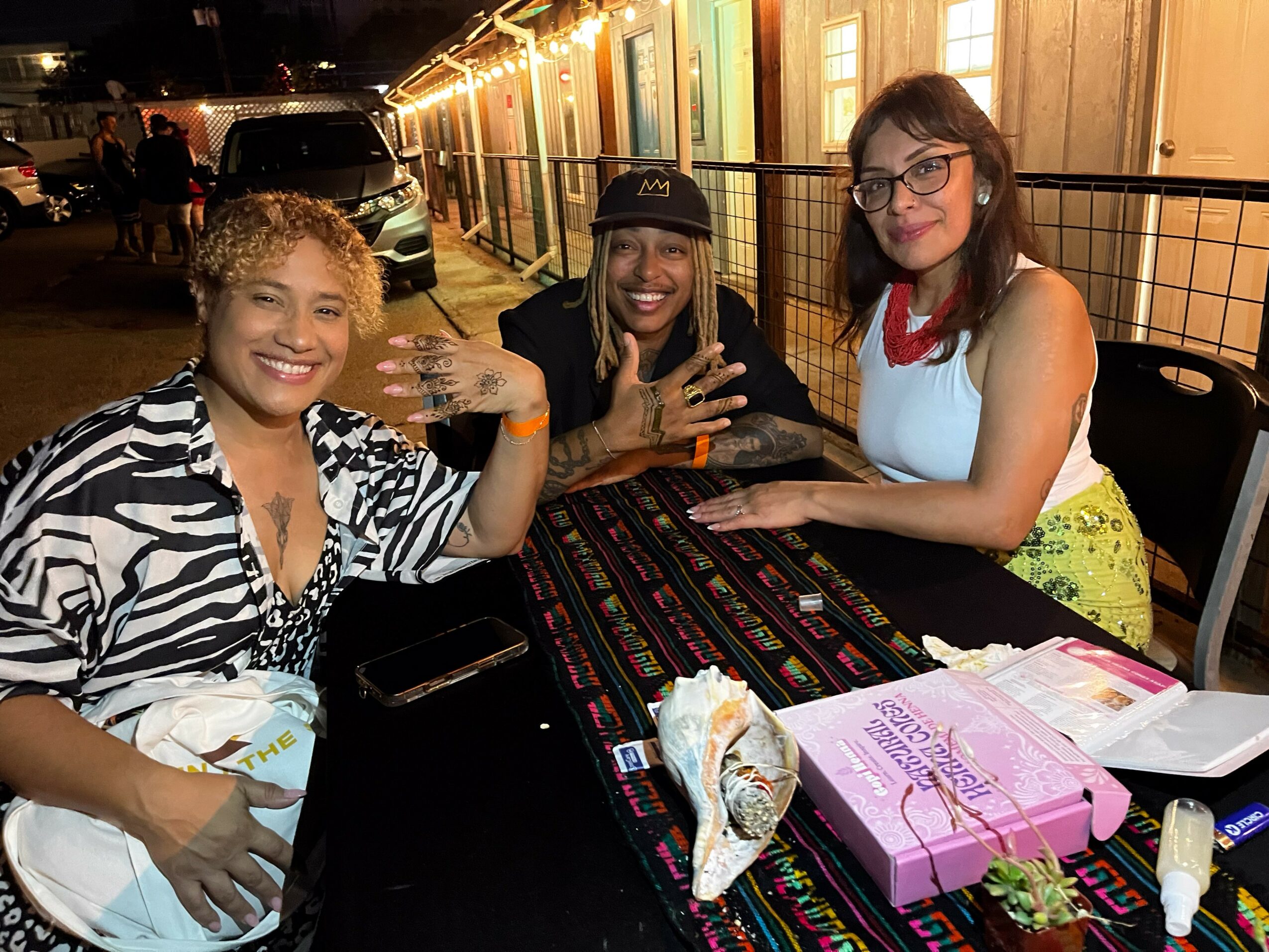 Three women sitting at a table outdoors at night, smiling, with a building and cars in the background.