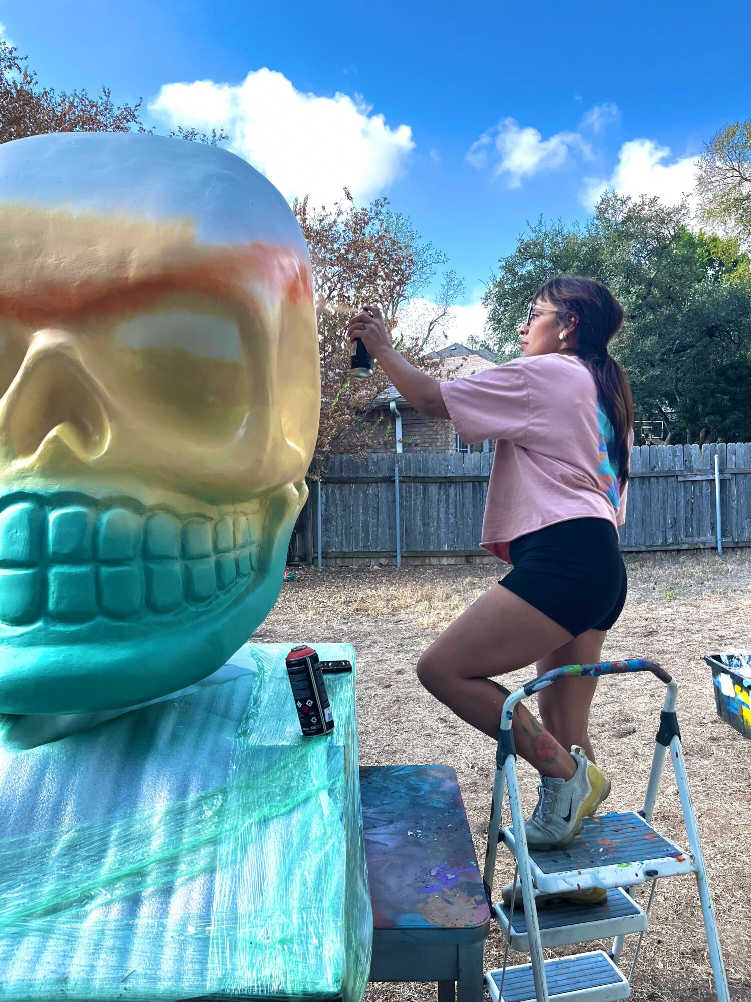 Person taking a photo outdoors, standing on a step ladder, with a large colorful skull sculpture nearby, trees, and a fence in the background.