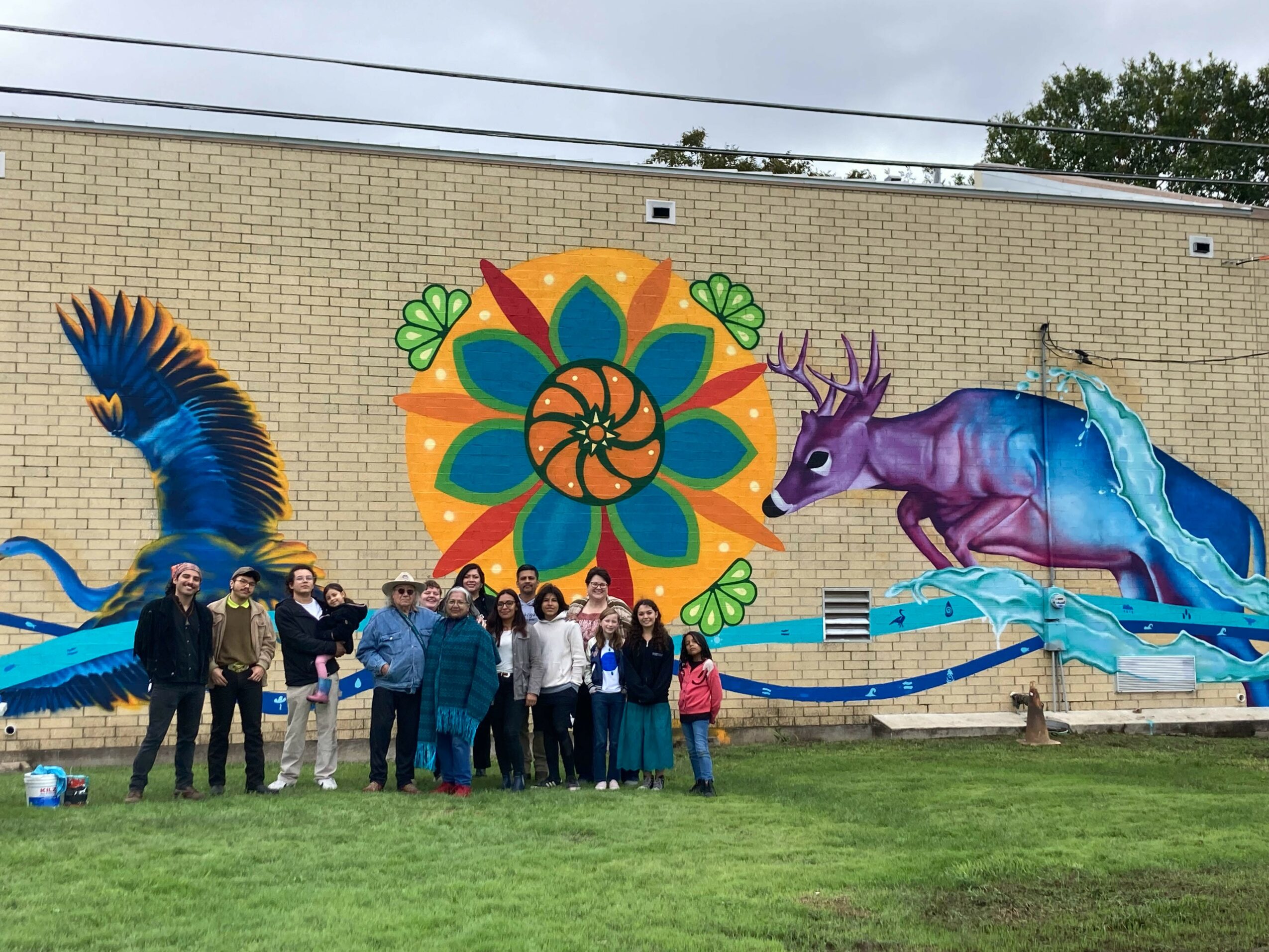 Group of people standing in front of a colorful mural with a bird, flower, and purple unicorn on a brick wall.
