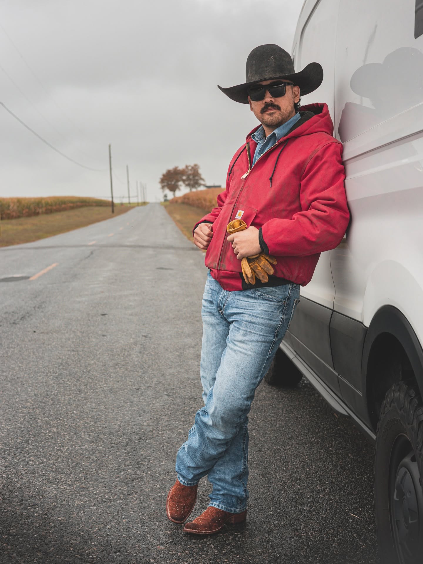Man leaning against a white vehicle on a rural road, wearing a cowboy hat, sunglasses, red jacket, and jeans.