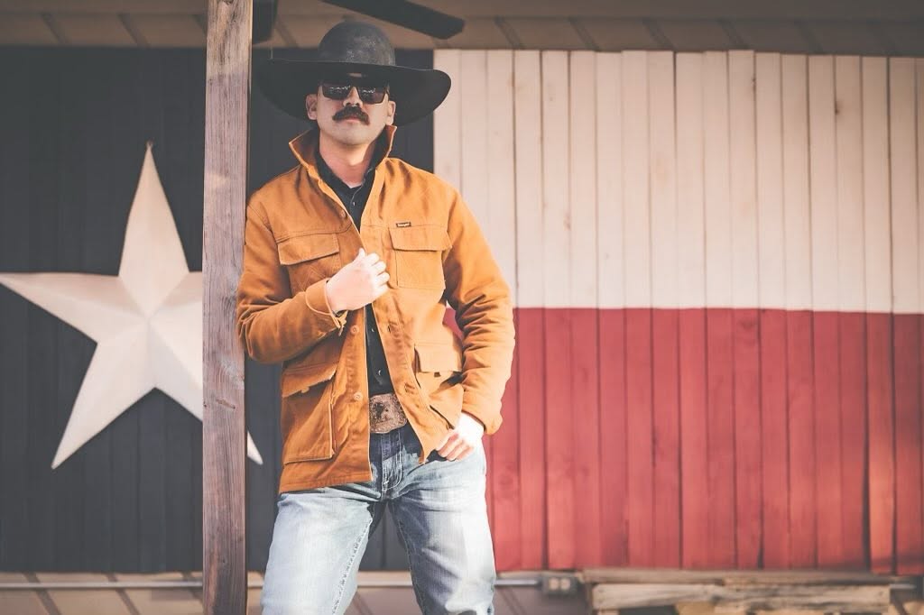 Man with a mustache wearing a wide-brimmed hat, orange jacket, and jeans, standing outdoors near a wooden post, with a star and striped background.