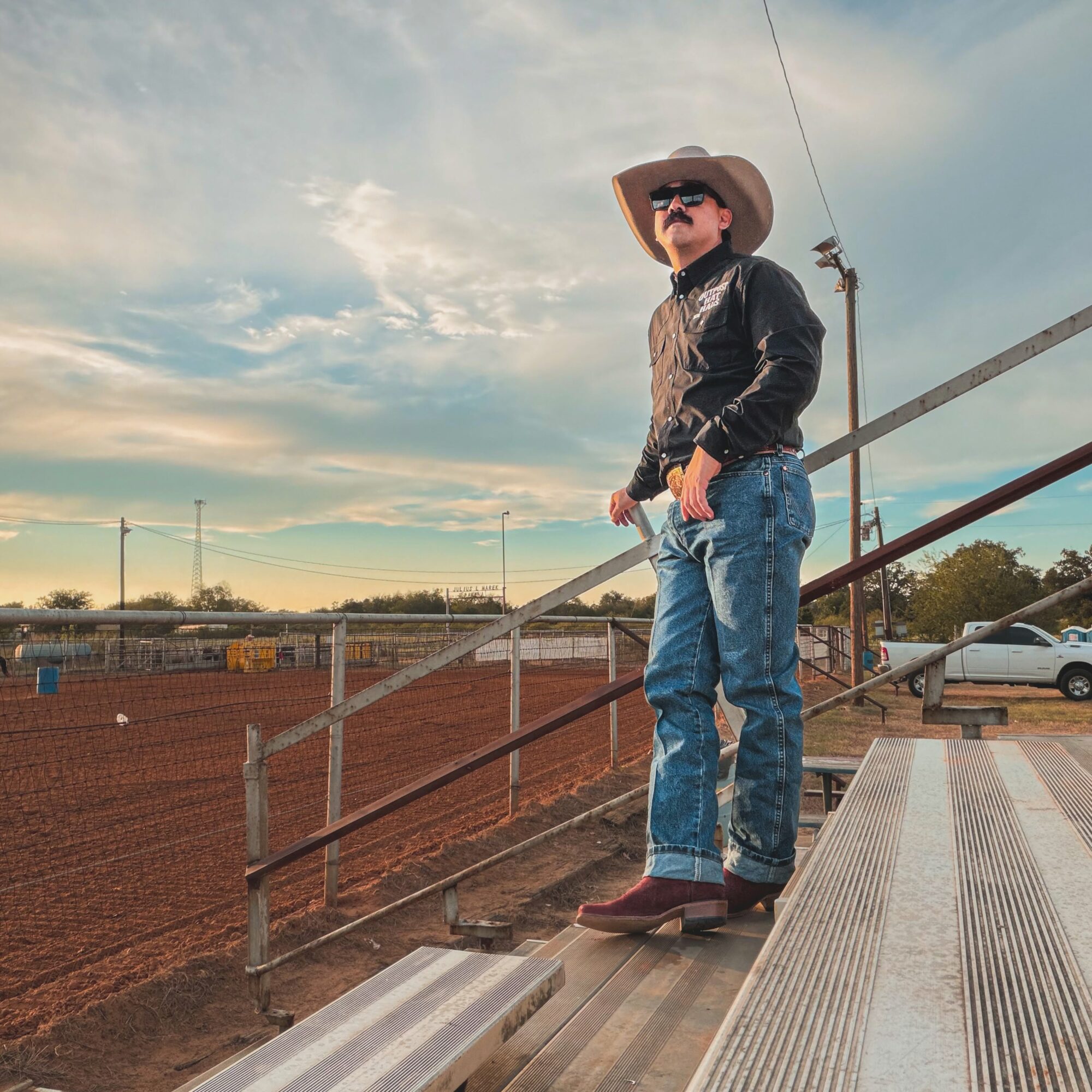 Person wearing a cowboy hat, sunglasses, leather jacket, and jeans standing on bleachers outdoors during daytime.