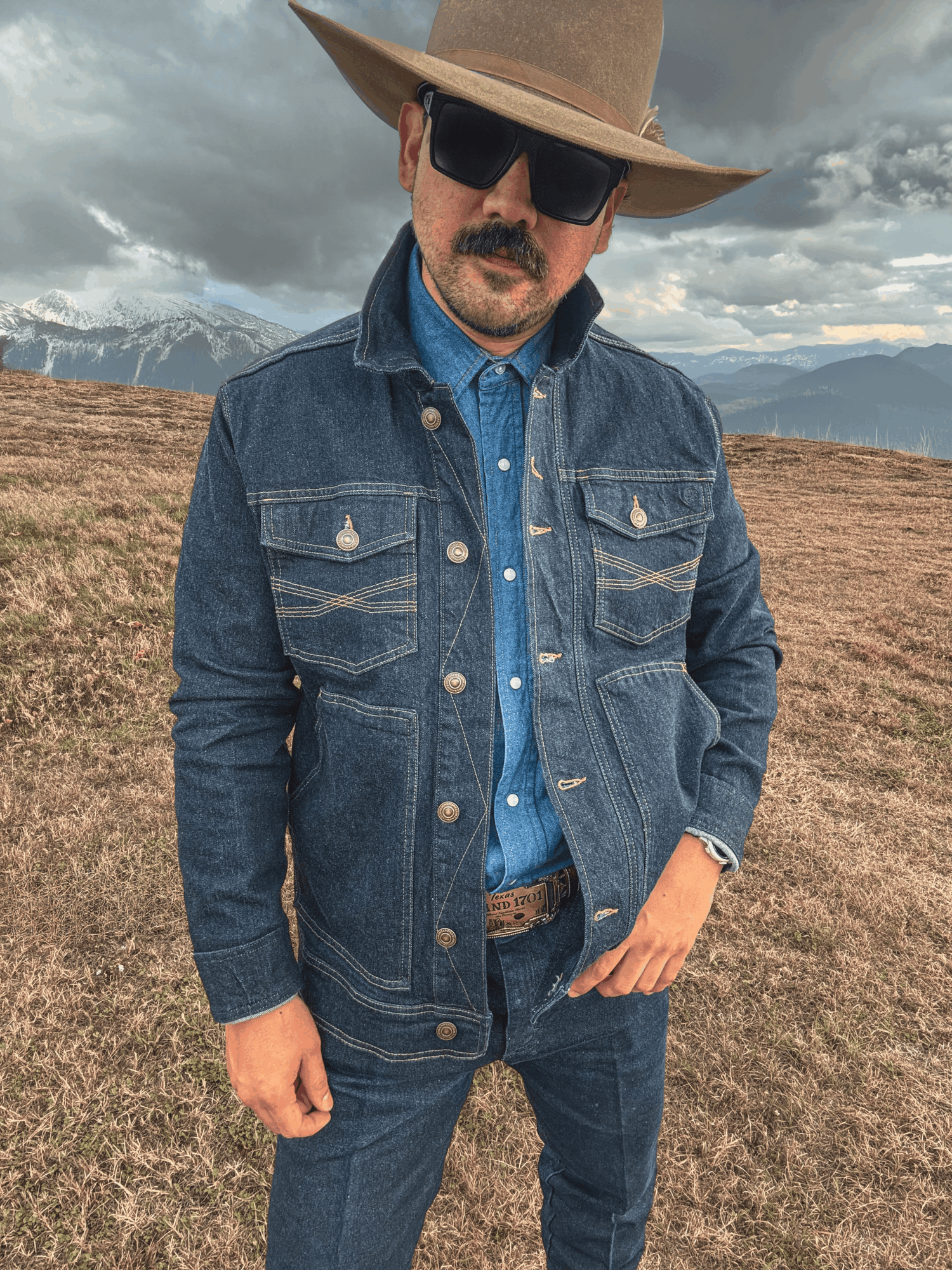 Man wearing a wide-brimmed hat, sunglasses, denim jacket, and jeans outdoors with mountains and cloudy sky in background.